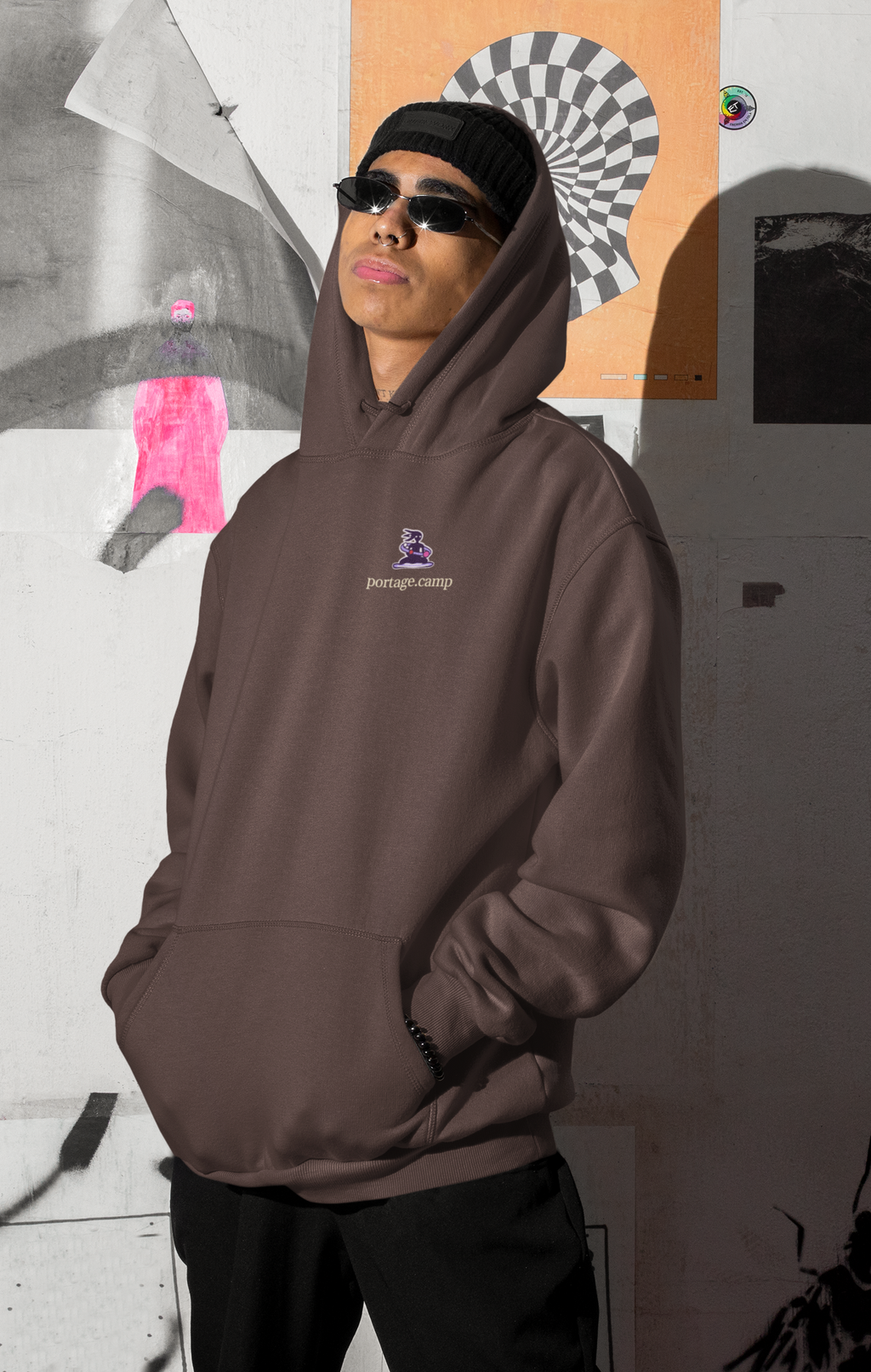 Orbit Keeper Heavyweight Hoodie in Brown, worn by a man by a wall of posters, with the Orbit Keeper logo and portage.camp on the left chest. Digital product mockup.