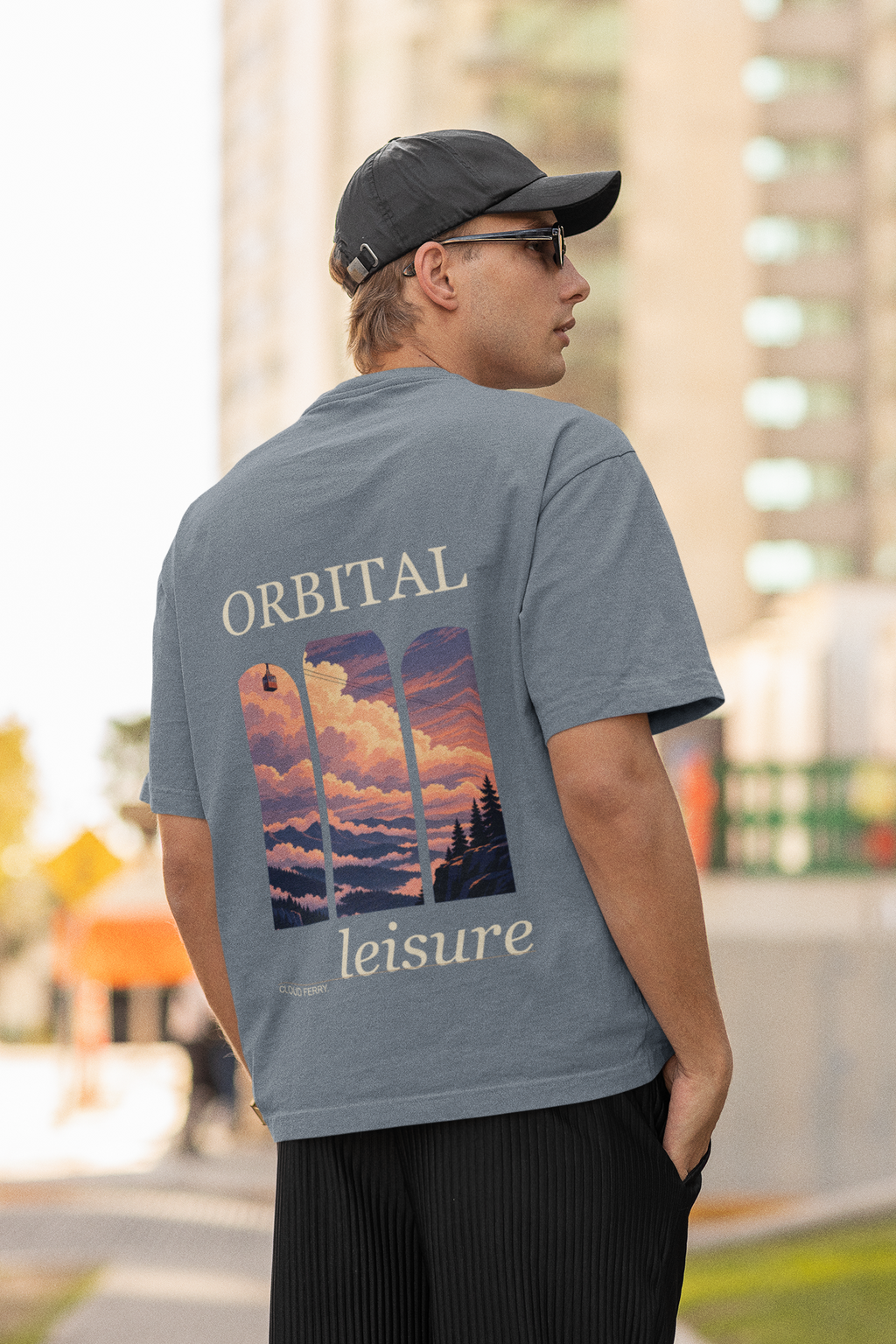 Cloud Ferry Garment-Dyed Tee in Blue Jean, worn by a man seen from behind in a cap and sunglasses on a city street, showing the large print on the back. Digital product mockup.