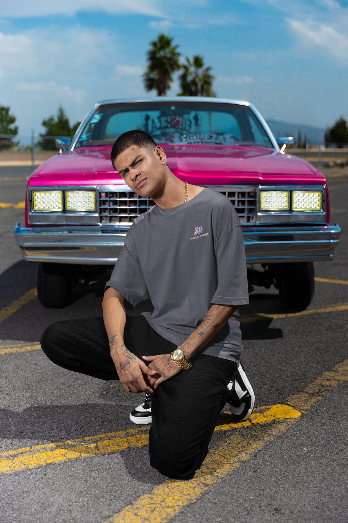 Amethyst After Hours Garment-Dyed Tee in Navy, worn by a man crouched in front of a pink classic car, with the Amethyst After Hours logo and portage.camp on the chest. Digital product mockup.