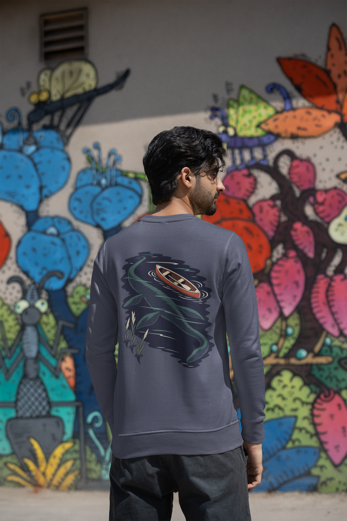 Seen Briefly Garment-Dyed Crewneck in Denim, worn by a man seen from behind by a painted wall in the city, showing the large print on the back. Digital product mockup.