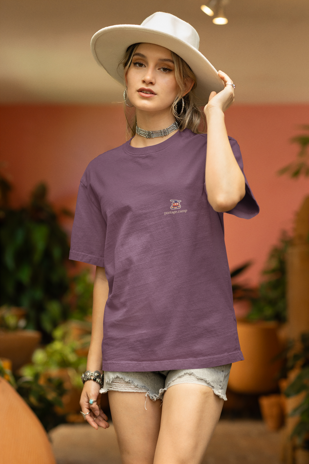 Cloud Ferry Garment-Dyed Tee in Berry, worn by a woman in a sun hat among potted plants, with the Cloud Ferry logo and portage.camp on the chest. Digital product mockup.
