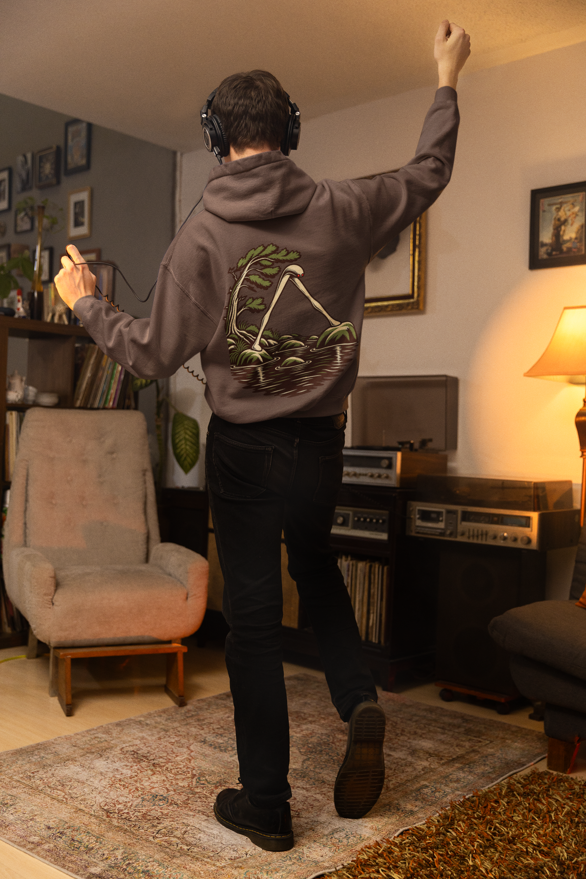 Small Hours Heavyweight Hoodie in Brown, worn by a man seen from behind in a living room at night, showing the large print on the back. Digital product mockup.