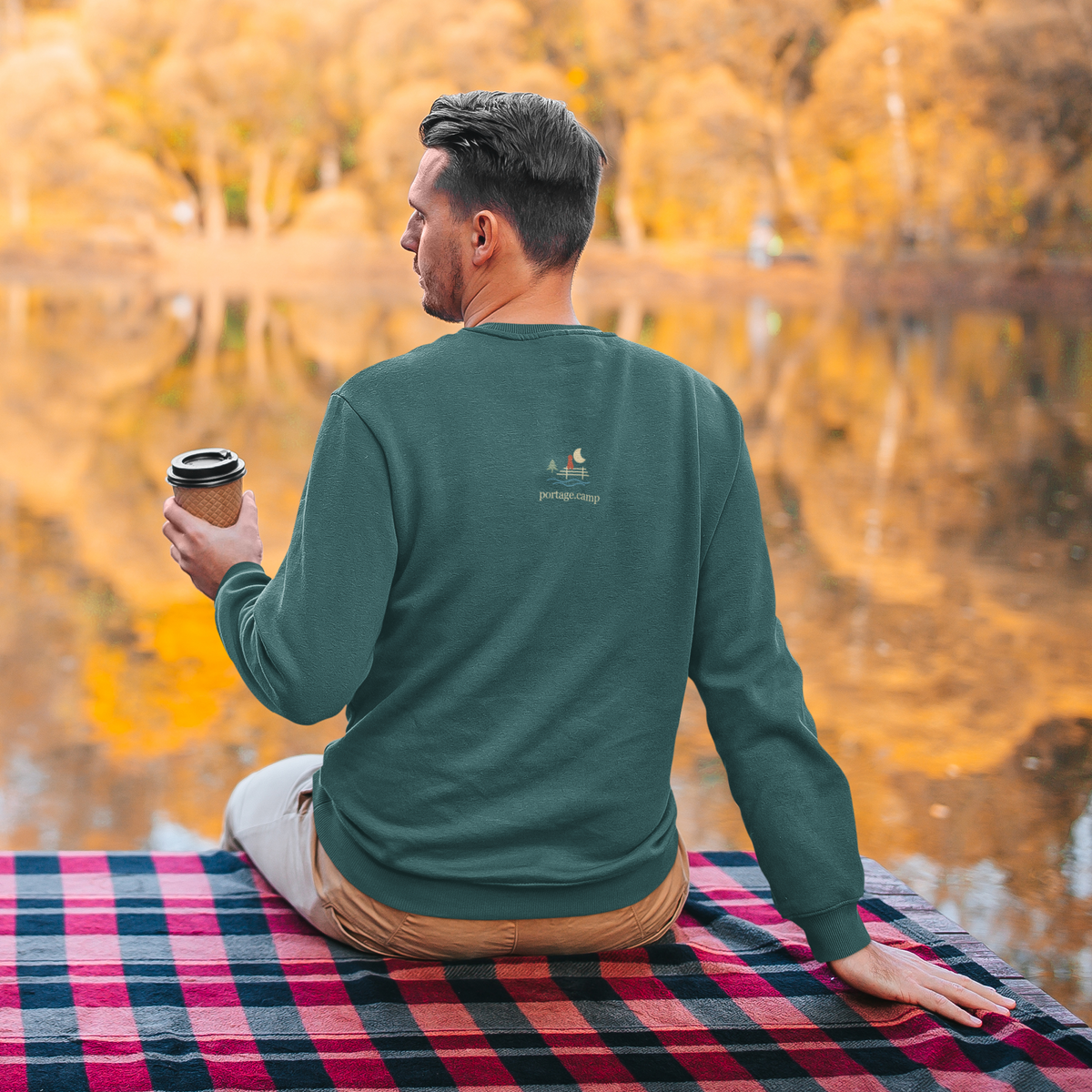 Quiet Hours Garment-Dyed Crewneck (Front Print) in Blue Spruce, worn by a man seen from behind seated by a lake in autumn, showing the portage.camp emblem at the upper back. Digital product mockup.