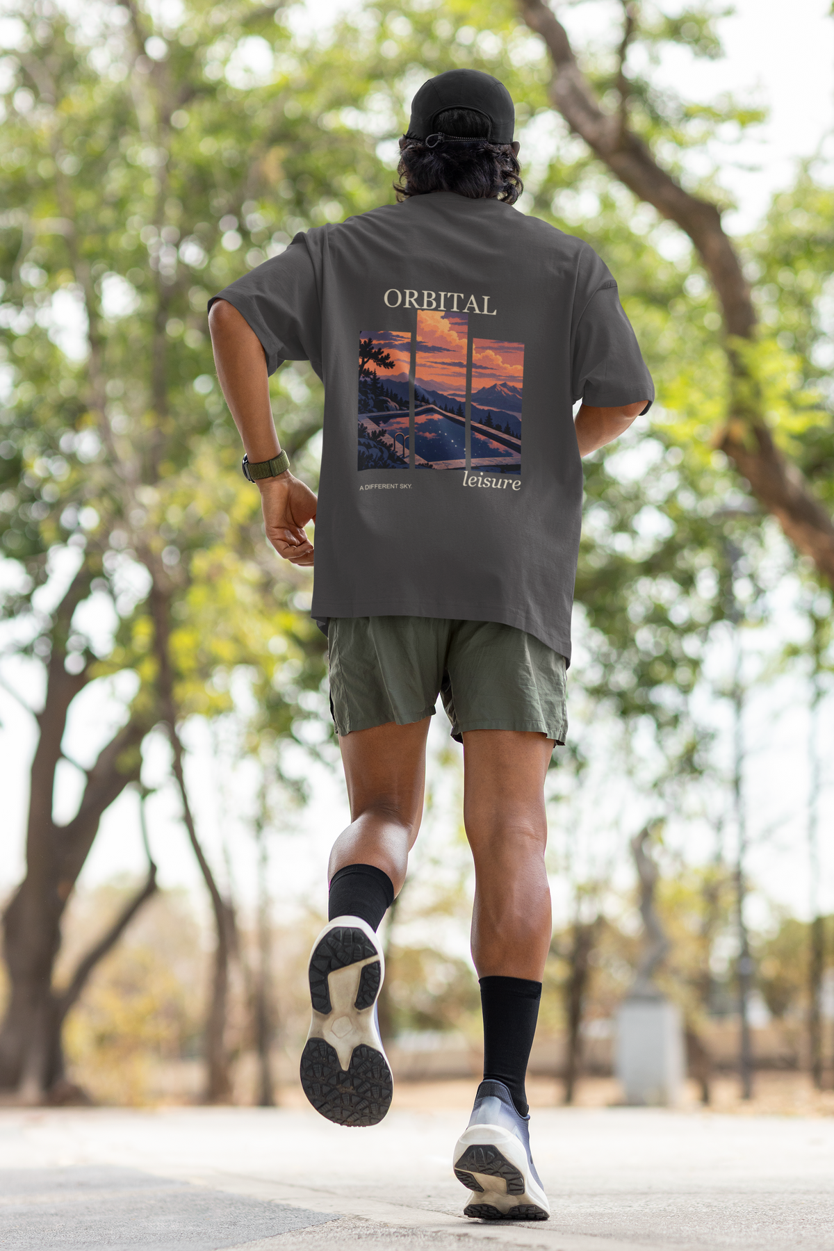 A Different Sky Garment-Dyed Tee in Graphite, worn by a man seen from behind jogging through a park, showing the large print on the back. Digital product mockup.