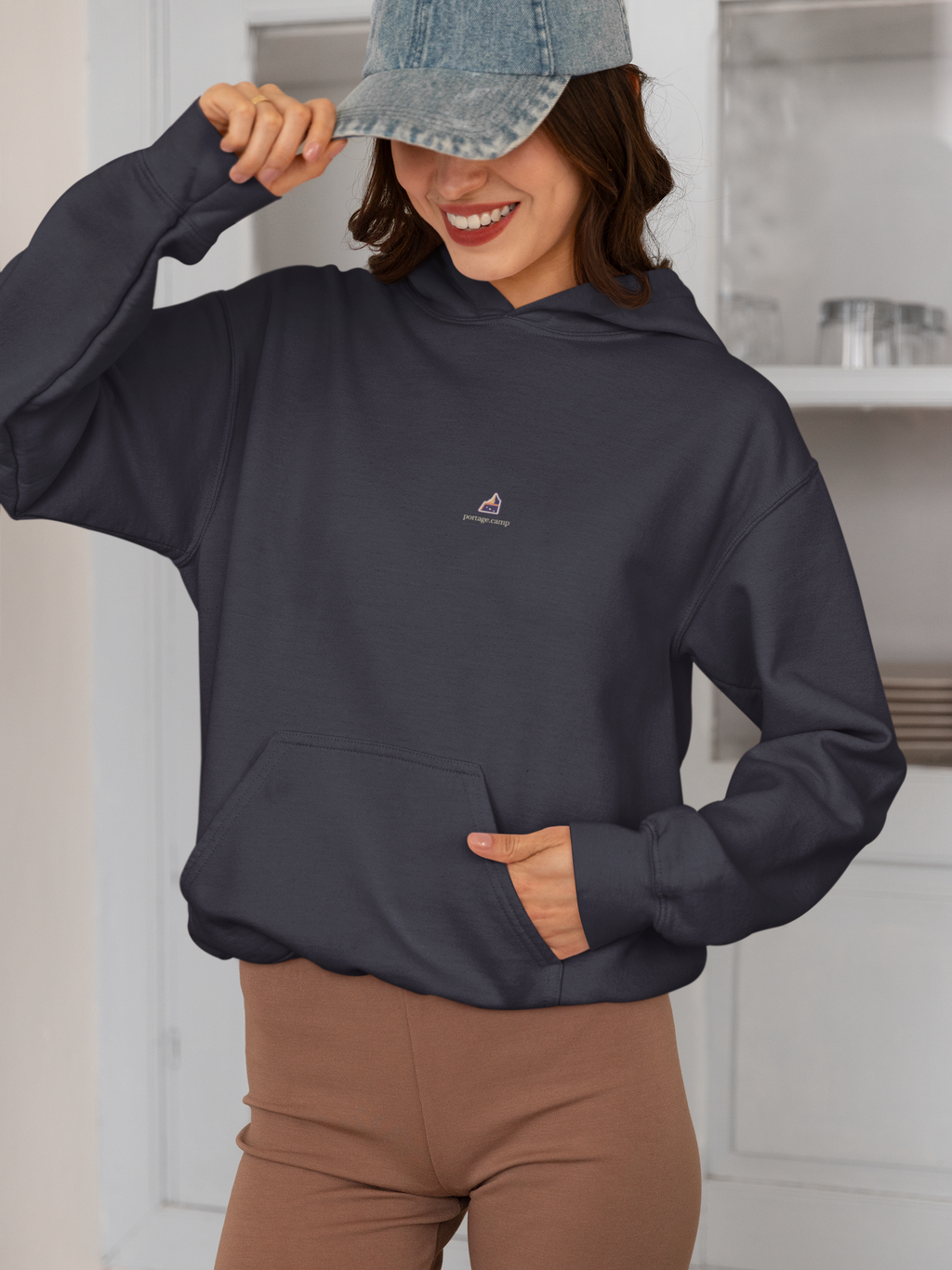 A Different Sky Midweight Hoodie in Classic Navy, worn by a woman smiling in a kitchen, with the A Different Sky logo and portage.camp on the chest. Digital product mockup.