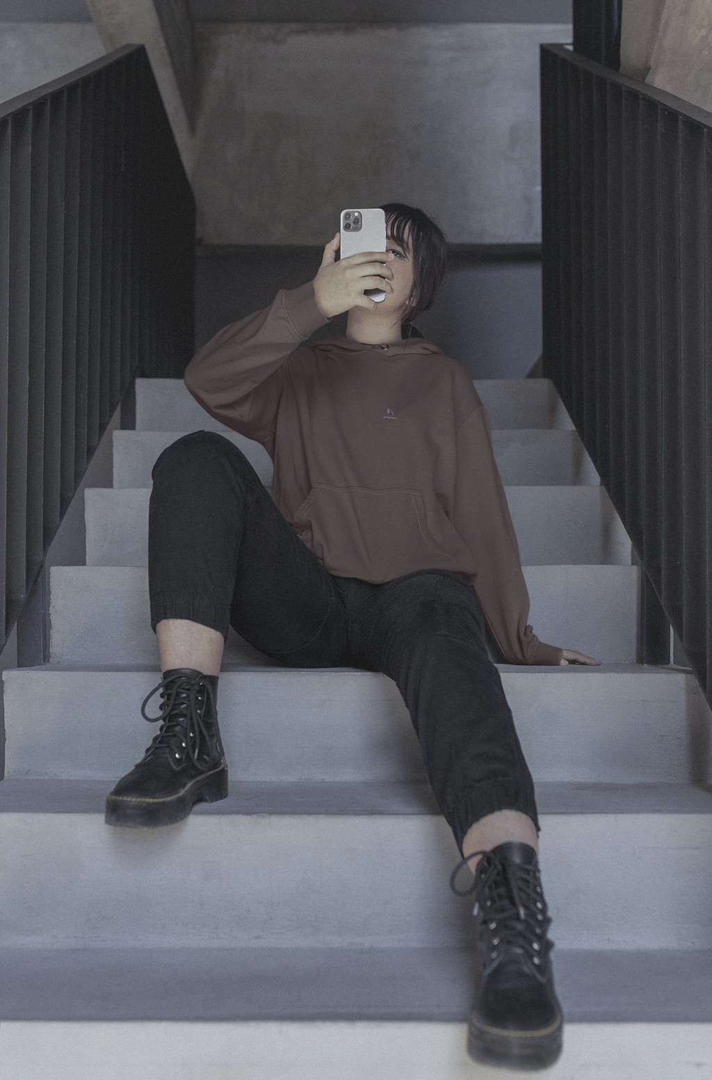 Orbit Keeper Heavyweight Hoodie in Brown, worn by a woman seated on a staircase, with the Orbit Keeper logo and portage.camp on the left chest. Digital product mockup.
