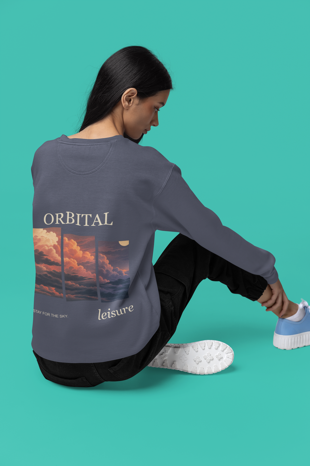 Stay for the Sky Garment-Dyed Crewneck in Denim, worn by a woman seen from behind seated on a studio floor, showing the large print on the back. Digital product mockup.