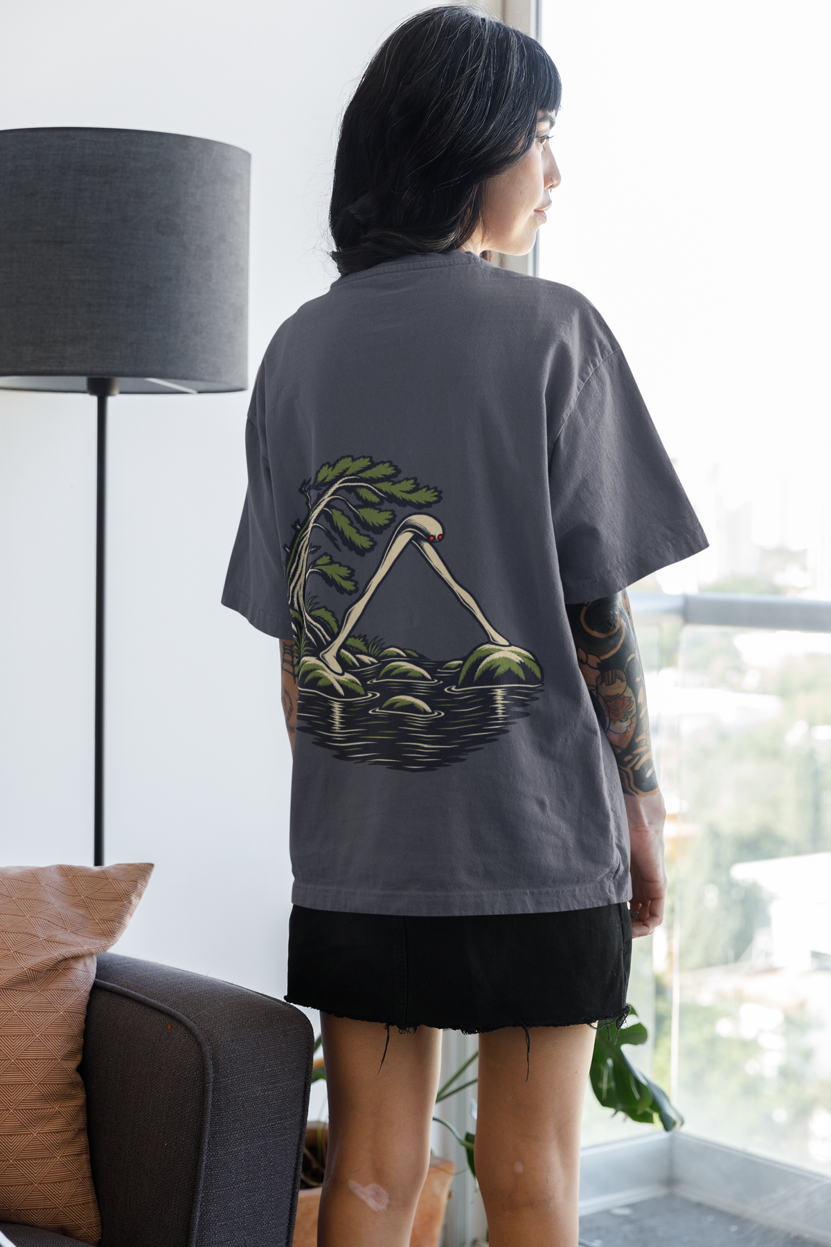 Small Hours Garment-Dyed Tee in Navy, worn by a woman seen from behind at home by a bright window, showing the large print on the back. Digital product mockup.