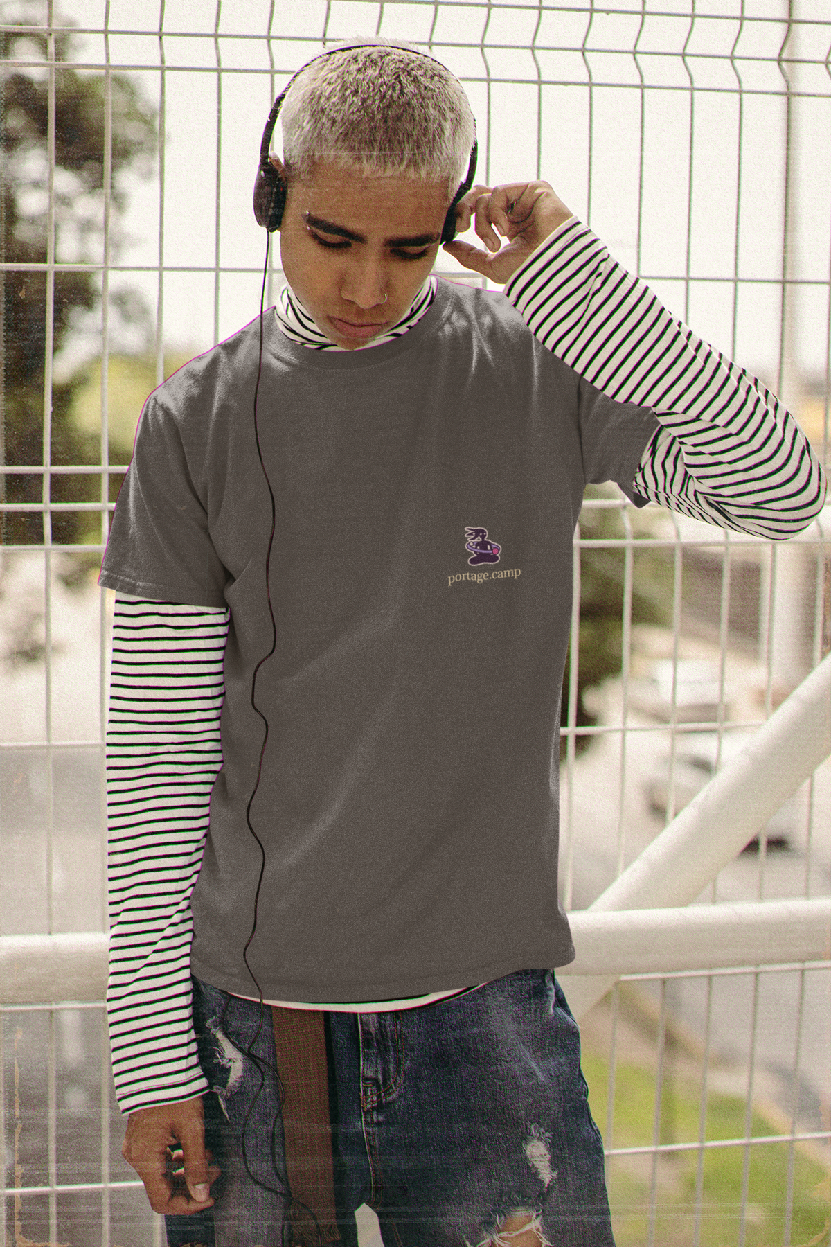 Orbit Keeper Garment-Dyed Tee in Graphite, worn by a man in headphones by a fence, with the Orbit Keeper logo and portage.camp on the chest. Digital product mockup.