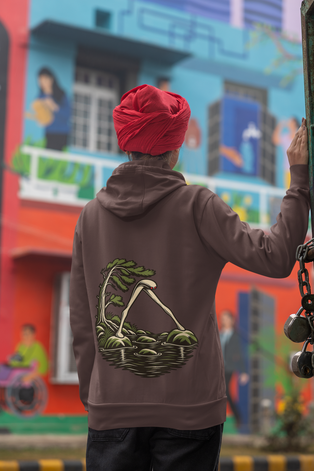 Open Late Heavyweight Hoodie in Brown, worn by a woman seen from behind in a turban by a painted mural, showing the large print on the back. Digital product mockup.