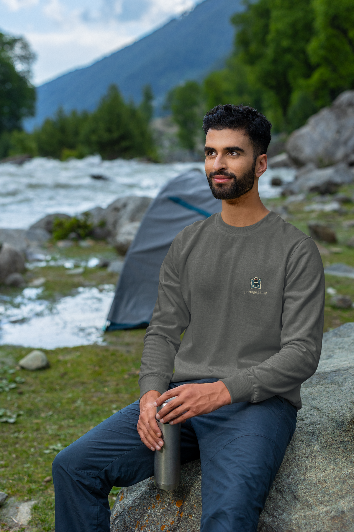 Night Library Garment-Dyed Crewneck in Pepper, worn by a man seated on a rock outdoors, with the Night Library logo and portage.camp on the chest. Digital product mockup.