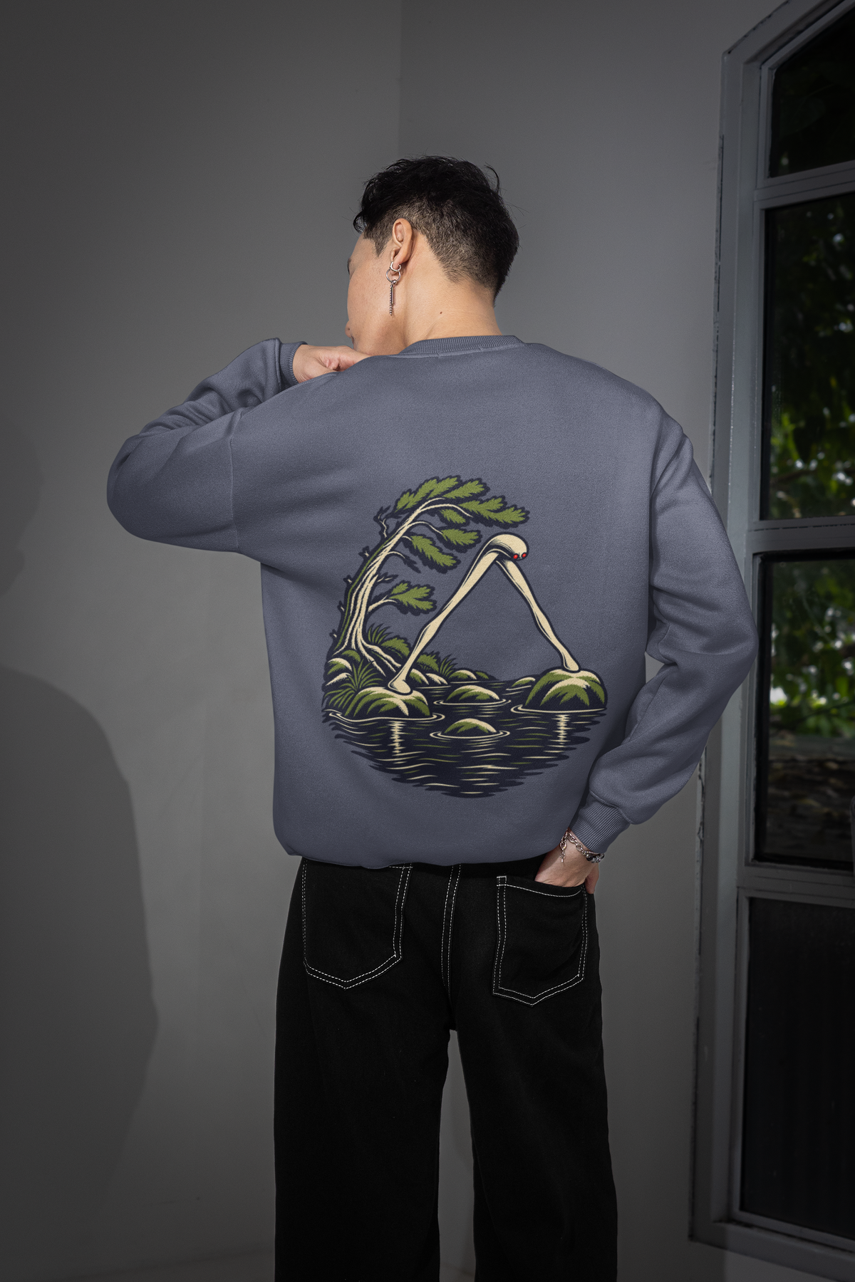 Small Hours Garment-Dyed Crewneck in Denim, worn by a man seen from behind in a room by a window, showing the large print on the back. Digital product mockup.