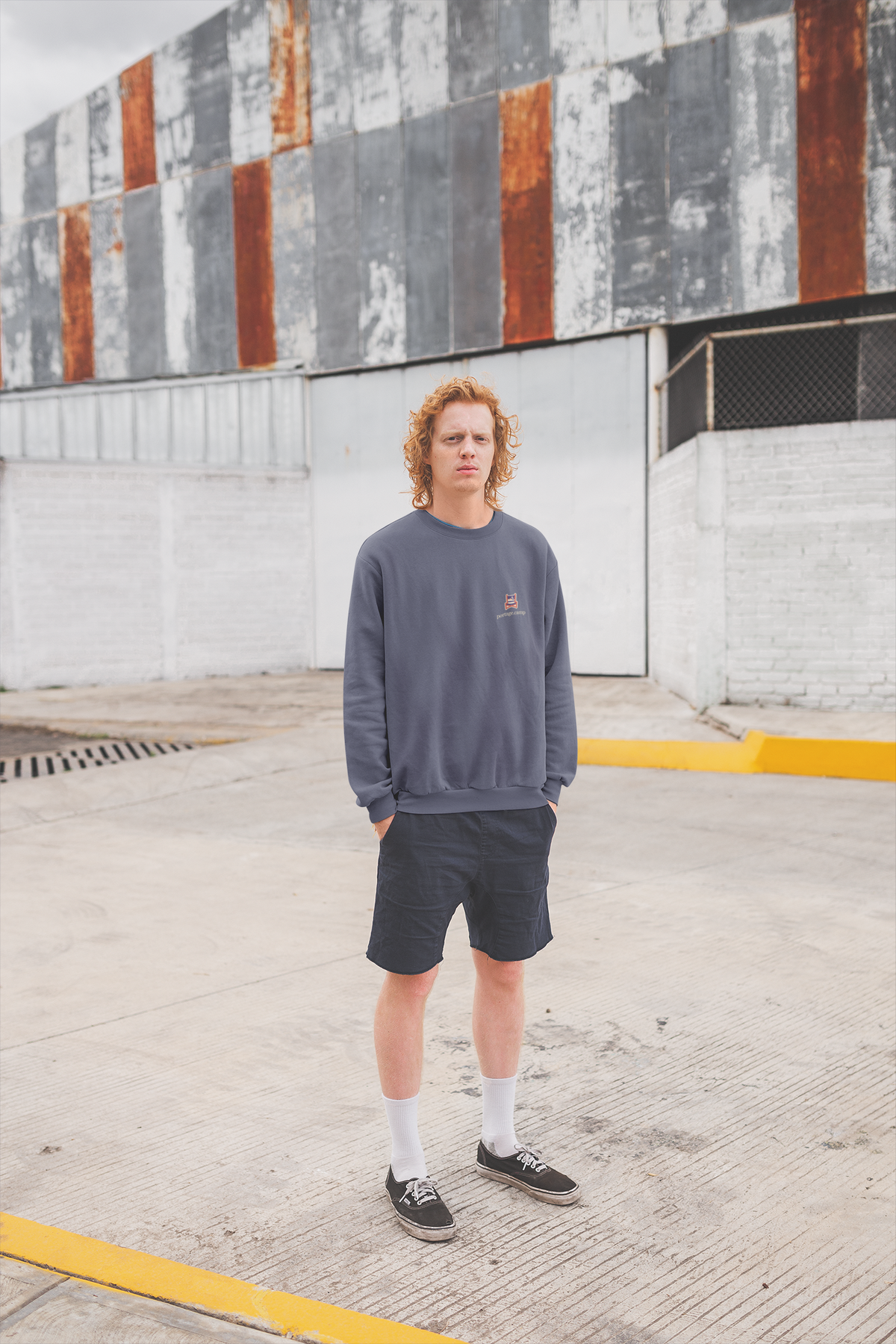 Edge of Evening Garment-Dyed Crewneck in Denim, worn by a man against a plain wall, with the Edge of Evening logo and portage.camp on the chest. Digital product mockup.