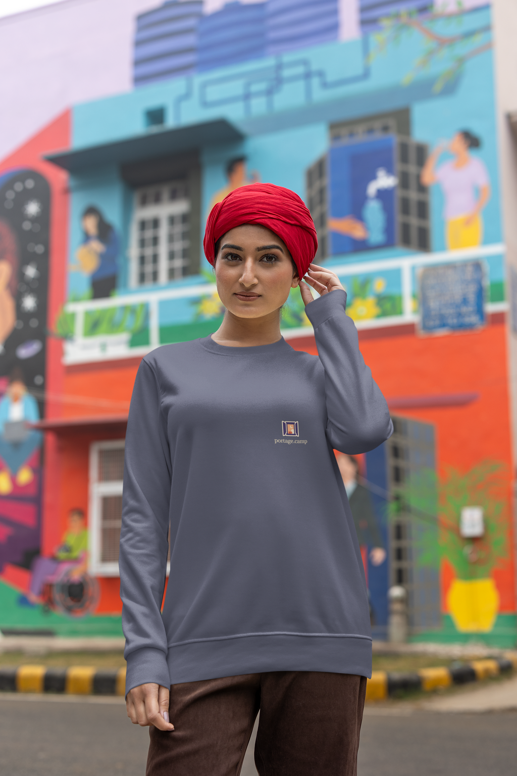 Open Late Garment-Dyed Crewneck in Denim, worn by a woman in a turban on a busy street, with the Open Late logo and portage.camp on the chest. Digital product mockup.