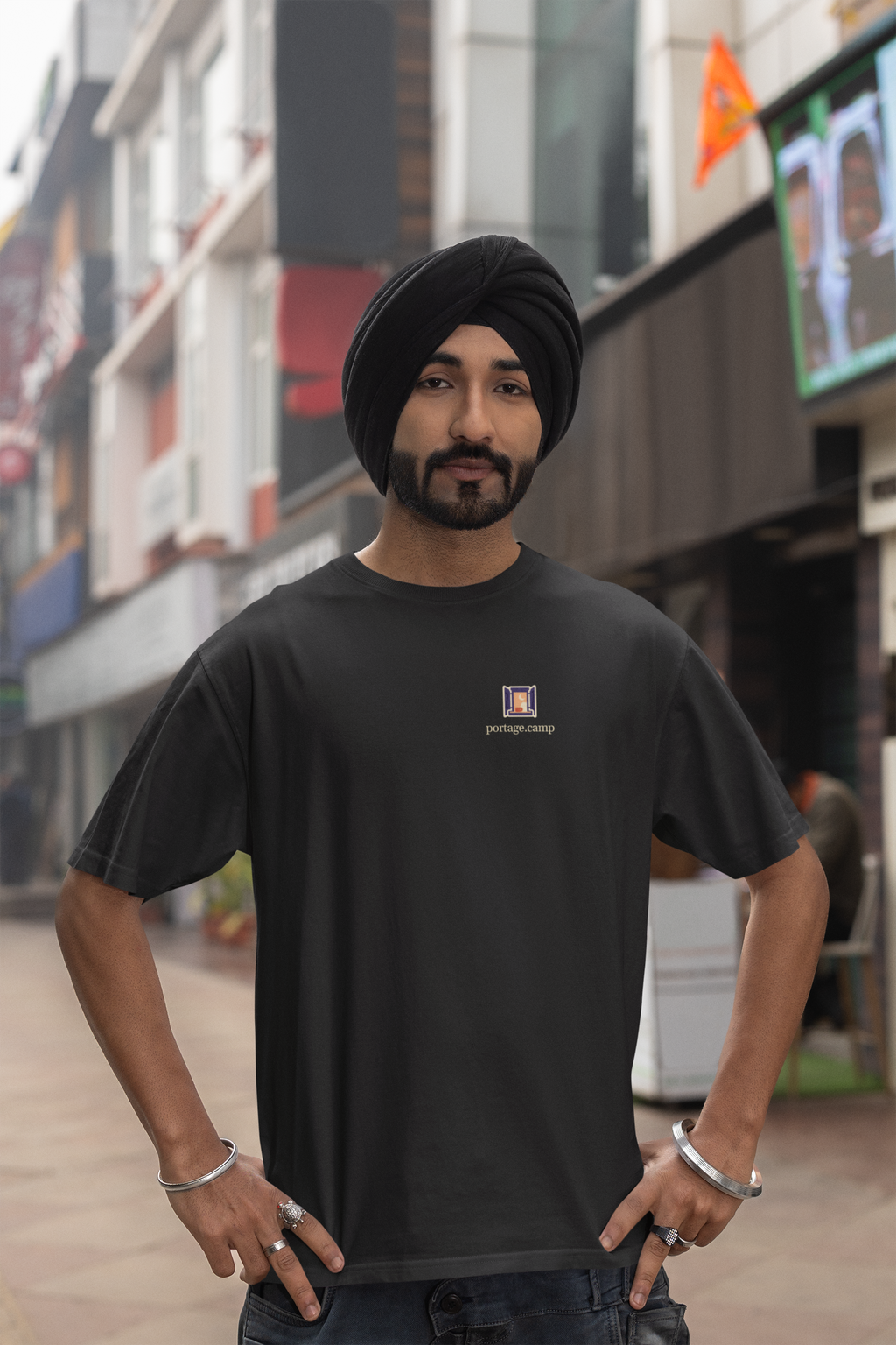 Open Late Garment-Dyed Tee in Black, worn by a man in a turban on a busy street, with the Open Late logo and portage.camp on the chest. Digital product mockup.
