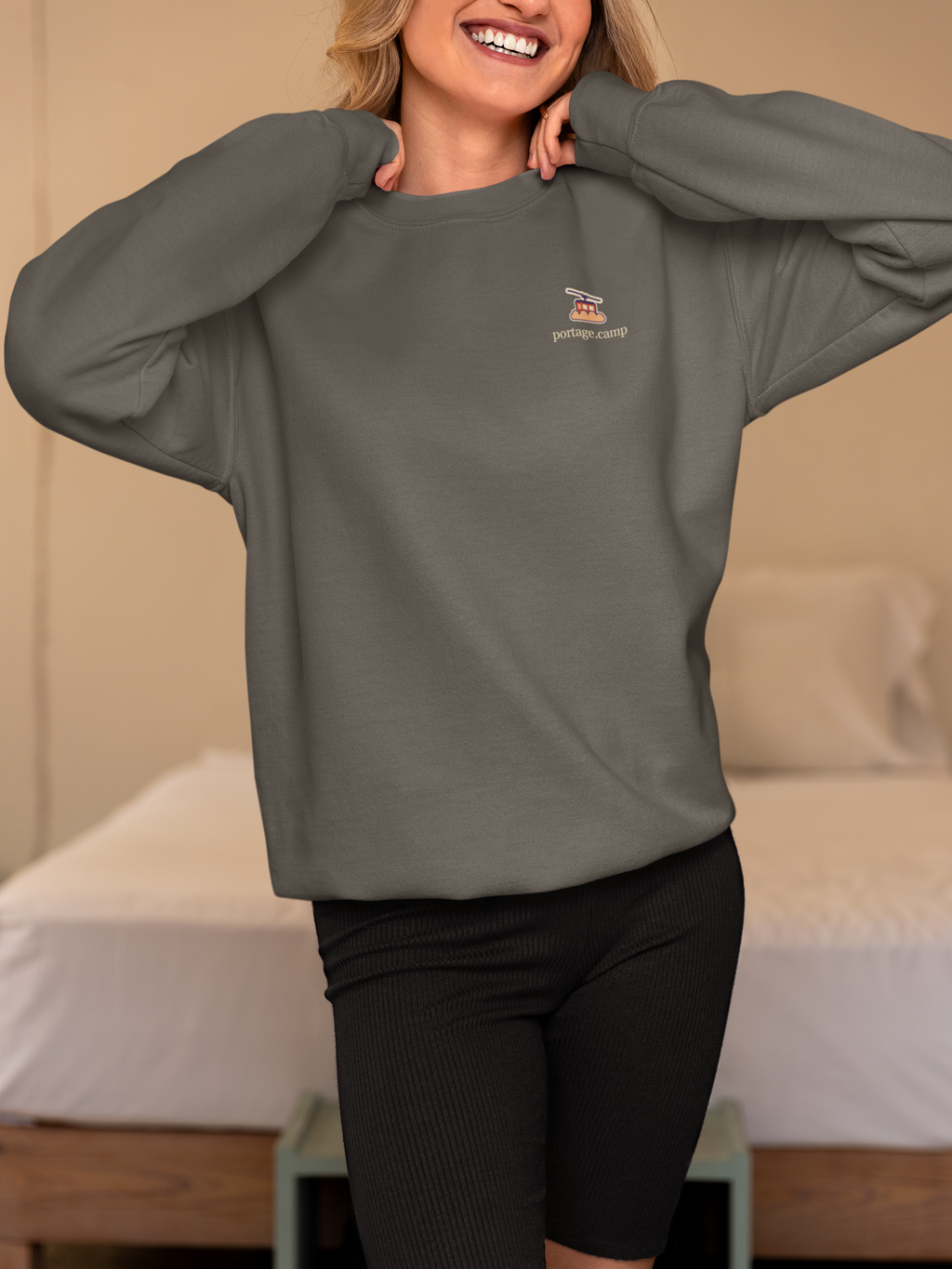 Cloud Ferry Garment-Dyed Crewneck in Pepper, worn by a woman in a bedroom, with the Cloud Ferry logo and portage.camp on the chest. Digital product mockup.