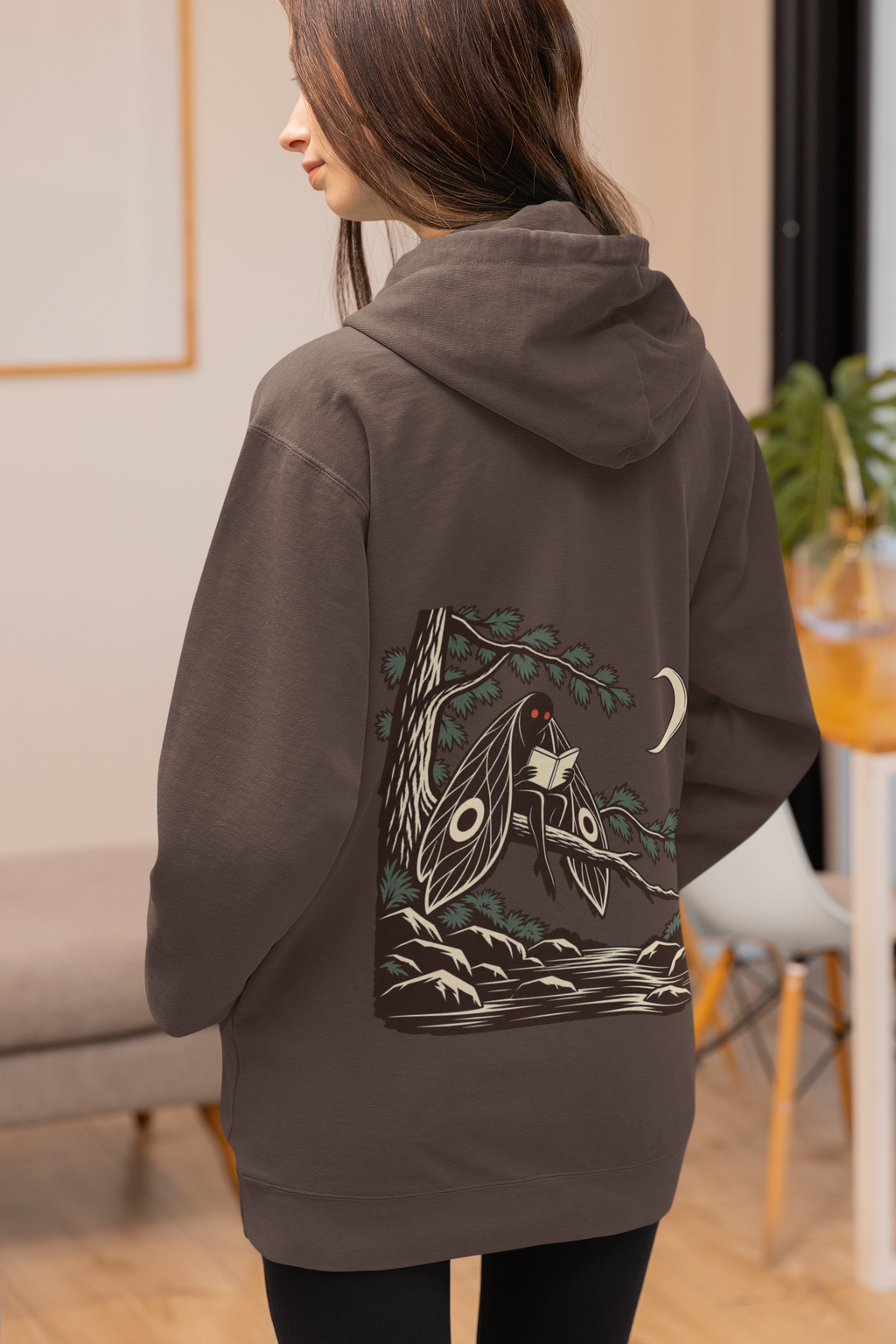 Night Library Heavyweight Hoodie in Brown, worn by a woman seen from behind at home, showing the large print on the back. Digital product mockup.