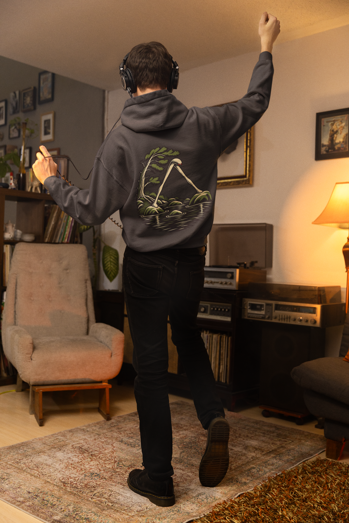 Small Hours Midweight Hoodie in Black, worn by a man seen from behind in a living room at night, showing the large print on the back. Digital product mockup.