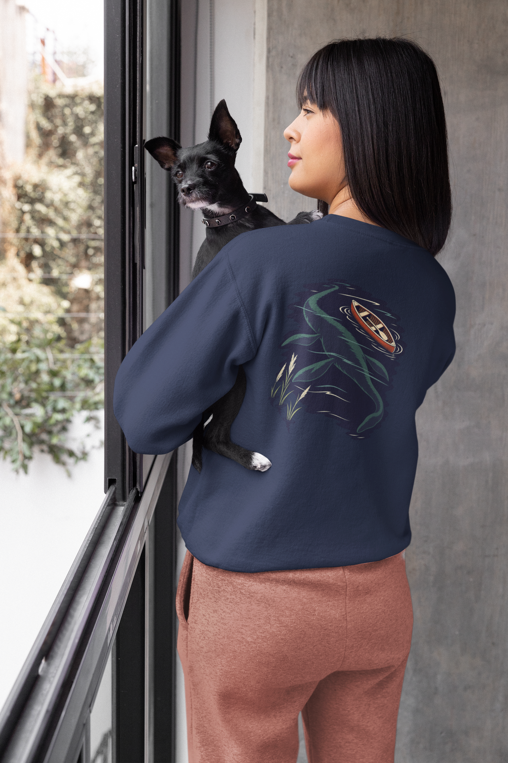 Seen Briefly Garment-Dyed Crewneck in True Navy, worn by a woman seen from behind at home, holding her dog, showing the large print on the back. Digital product mockup.