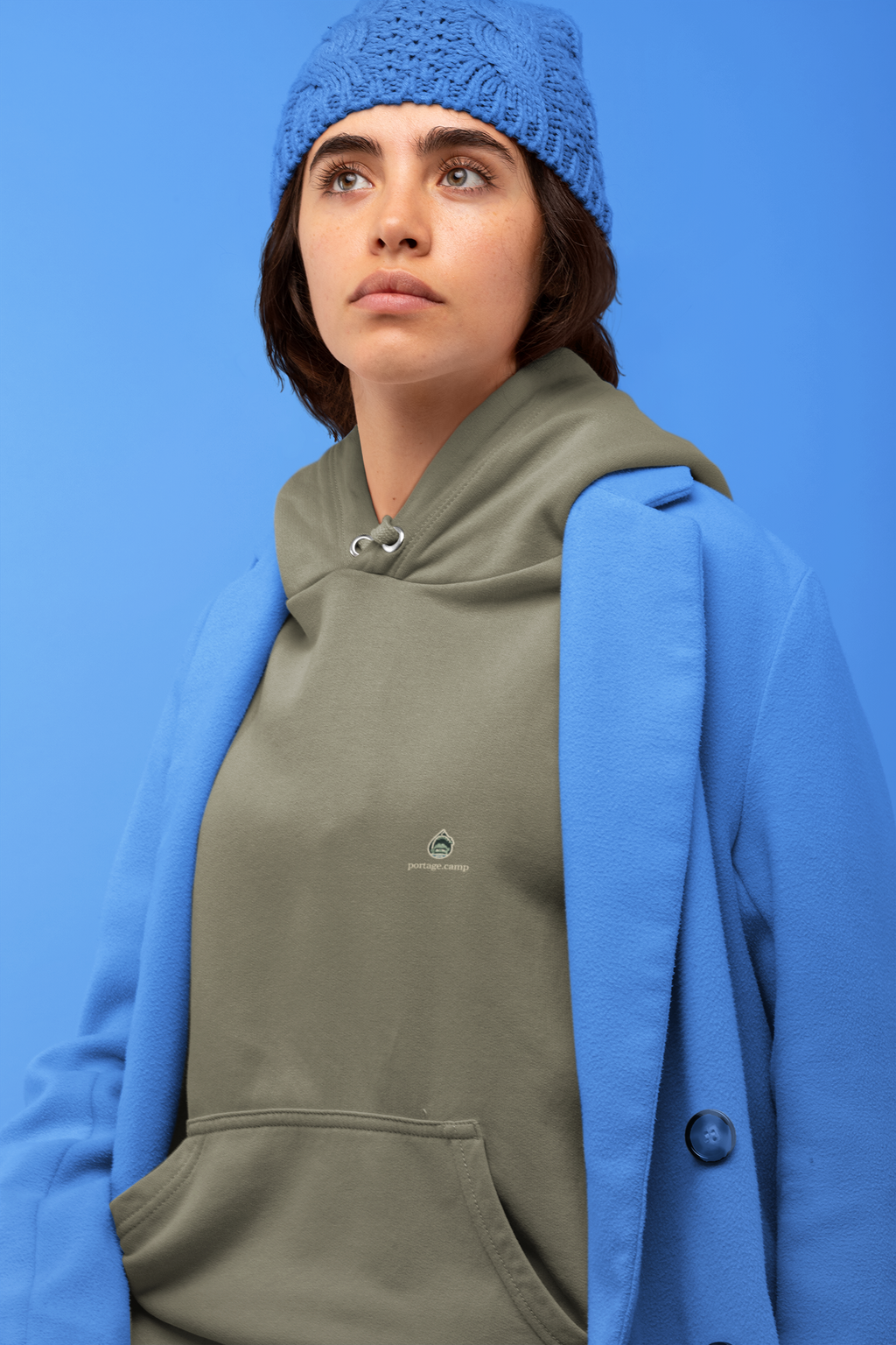 Rain Check Heavyweight Hoodie in Olive, worn by a woman against a blue backdrop, with the Rain Check logo and portage.camp on the left chest. Digital product mockup.