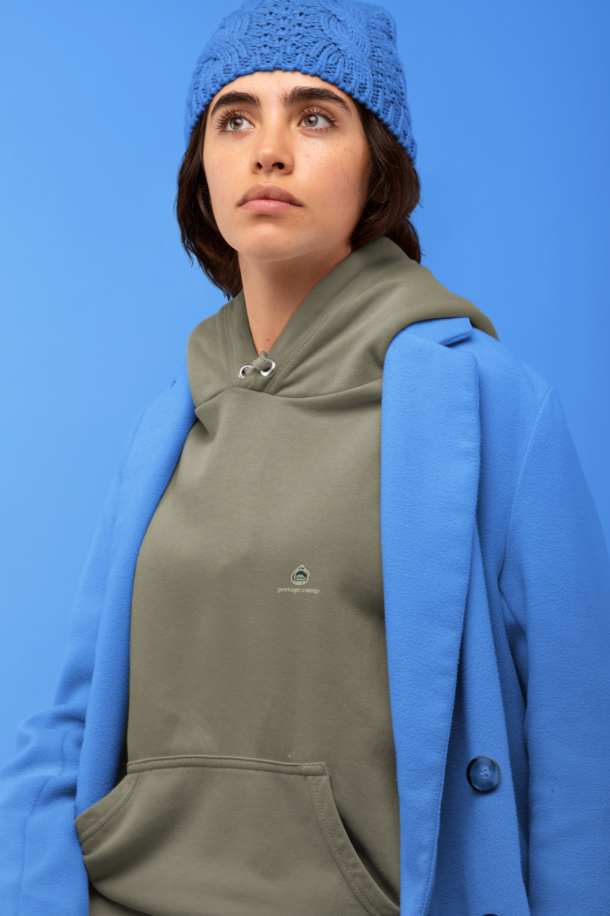 Rain Check Heavyweight Hoodie in Olive, worn by a woman against a blue backdrop, with the Rain Check logo and portage.camp on the left chest. Digital product mockup.