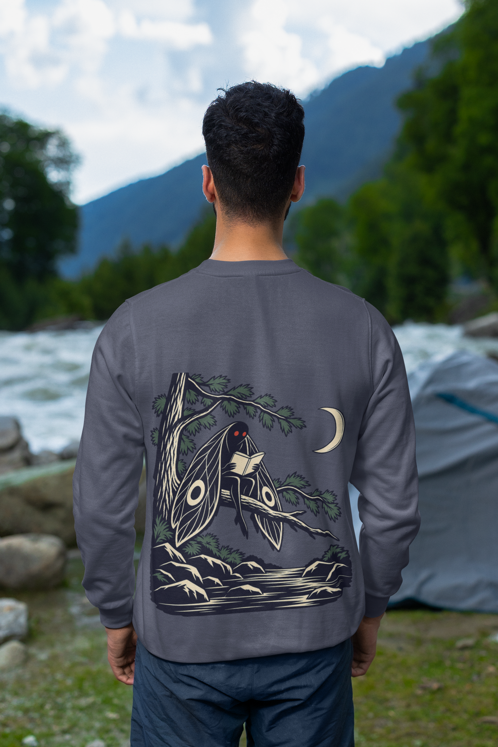 Night Library Garment-Dyed Crewneck in Denim, worn by a man seen from behind looking out over a mountain valley, showing the large print on the back. Digital product mockup.