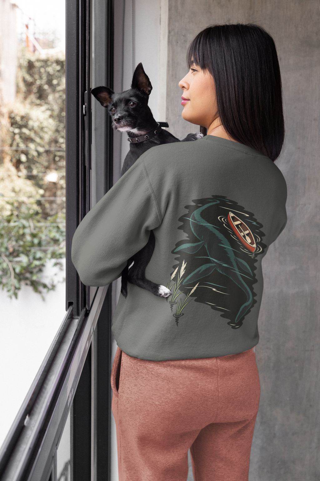 Seen Briefly Garment-Dyed Crewneck in Pepper, worn by a woman seen from behind at home, holding her dog, showing the large print on the back. Digital product mockup.