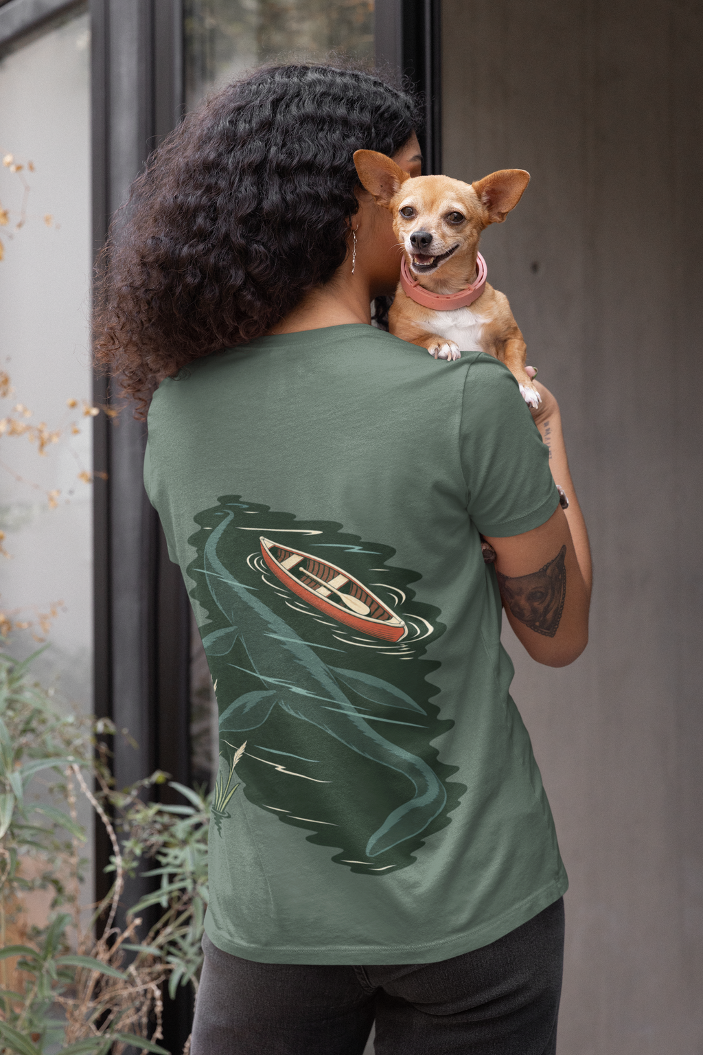 Seen Briefly Garment-Dyed Tee in Blue Spruce, worn by a woman seen from behind holding a small dog, showing the large print on the back. Digital product mockup.