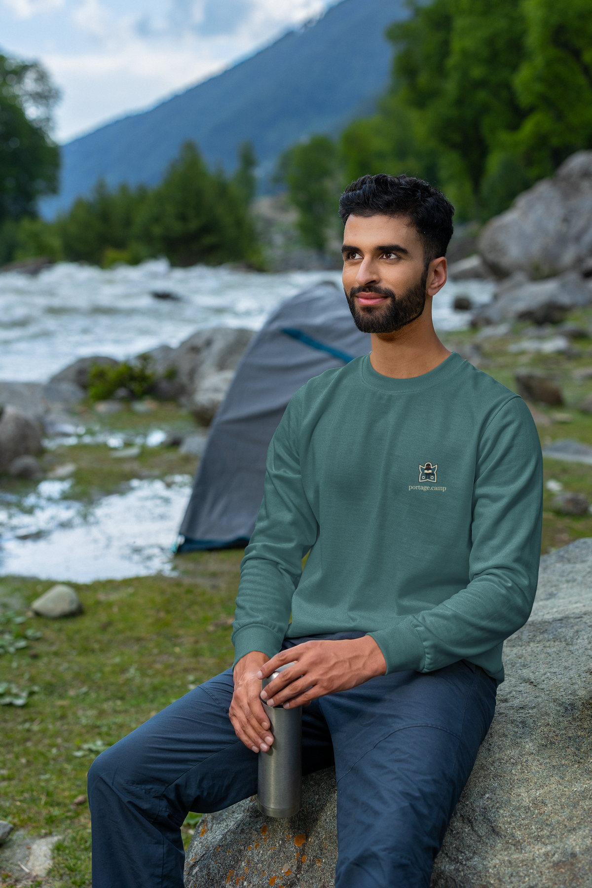 Night Library Garment-Dyed Crewneck in Blue Spruce, worn by a man seated on a rock outdoors, with the Night Library logo and portage.camp on the chest. Digital product mockup.