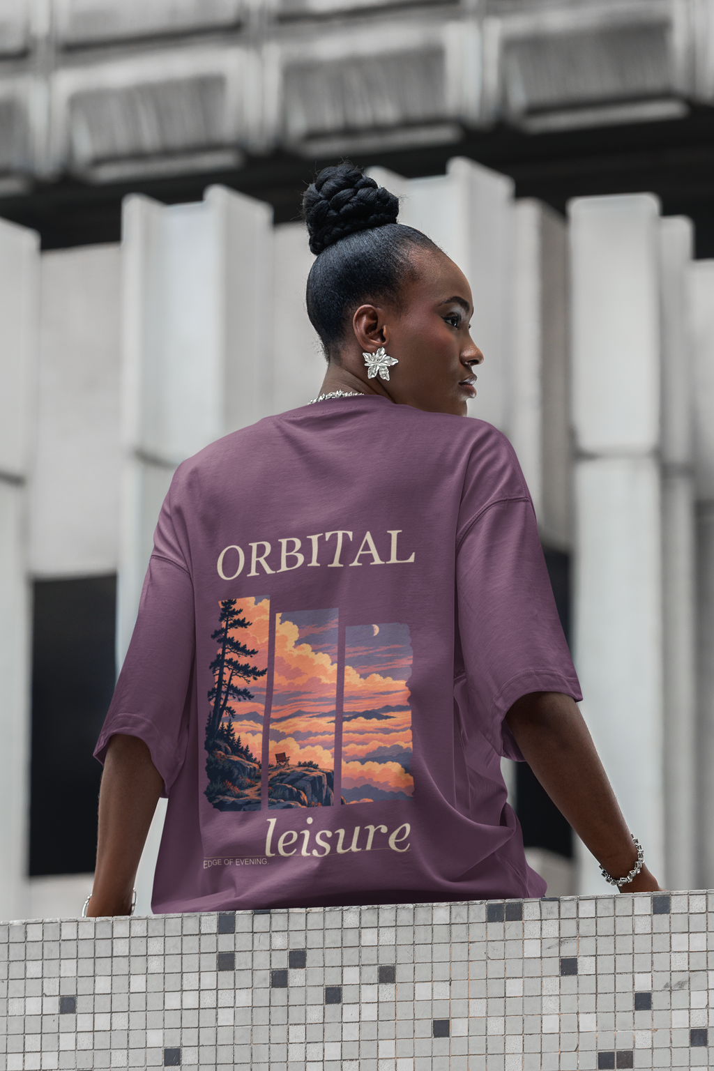 Edge of Evening Garment-Dyed Tee in Berry, worn by a woman seen from behind on a city street lined with columns, showing the large print on the back. Digital product mockup.