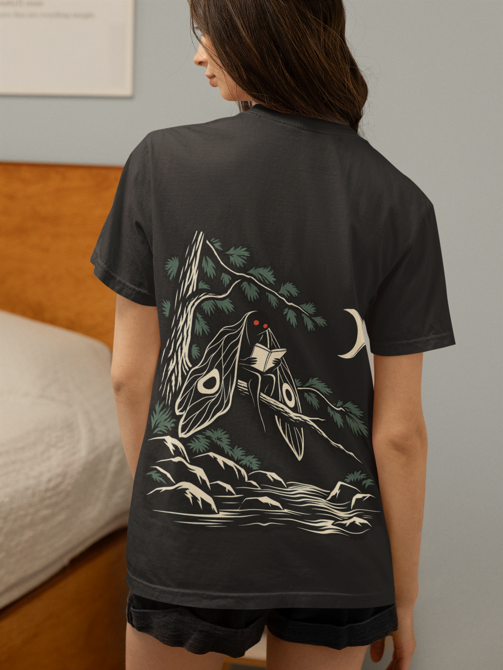 Night Library Garment-Dyed Tee in Black, worn by a woman seen from behind in a bedroom at home, showing the large print on the back. Digital product mockup.