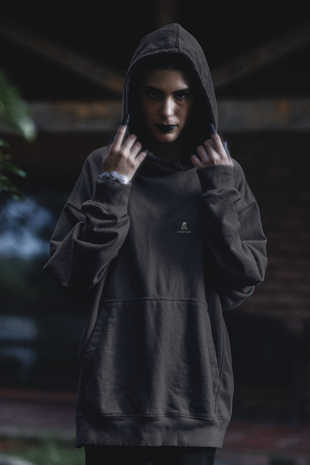 Small Hours Heavyweight Hoodie in Black, worn by a woman in a dark forest, with the Small Hours logo and portage.camp on the left chest. Digital product mockup.