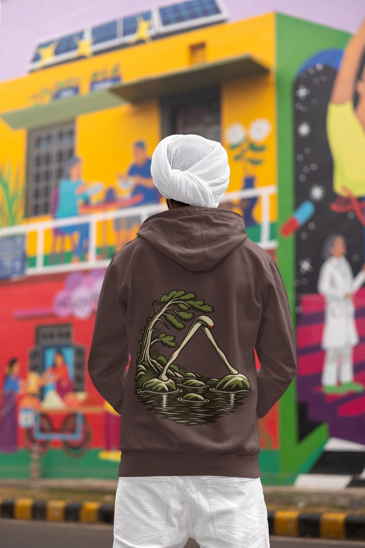Open Late Heavyweight Hoodie in Brown, worn by a man seen from behind in a turban by a painted mural, showing the large print on the back. Digital product mockup.