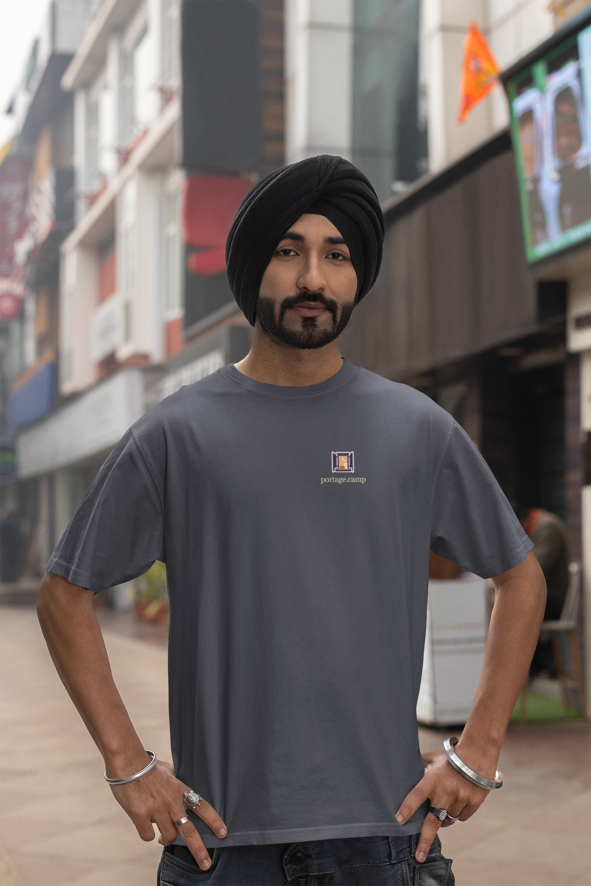Open Late Garment-Dyed Tee in Navy, worn by a man in a turban on a busy street, with the Open Late logo and portage.camp on the chest. Digital product mockup.