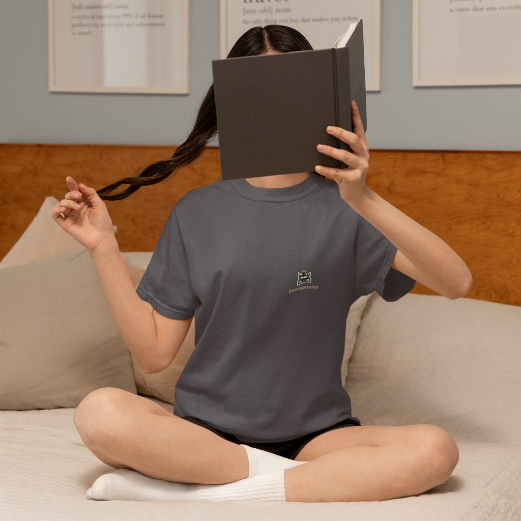 Night Library Garment-Dyed Tee in Navy, worn by a woman at home, hiding behind a book, with the Night Library logo and portage.camp on the chest. Digital product mockup.