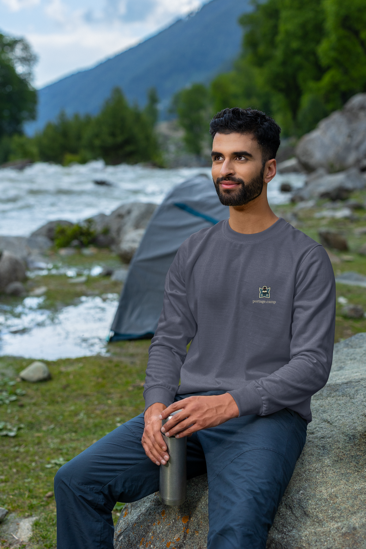 Night Library Garment-Dyed Crewneck in Denim, worn by a man seated on a rock outdoors, with the Night Library logo and portage.camp on the chest. Digital product mockup.