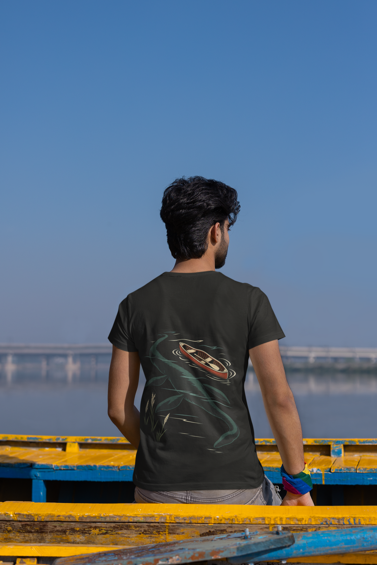 Seen Briefly Garment-Dyed Tee in Black, worn by a man seen from behind seated on a canoe by the water, showing the large print on the back. Digital product mockup.