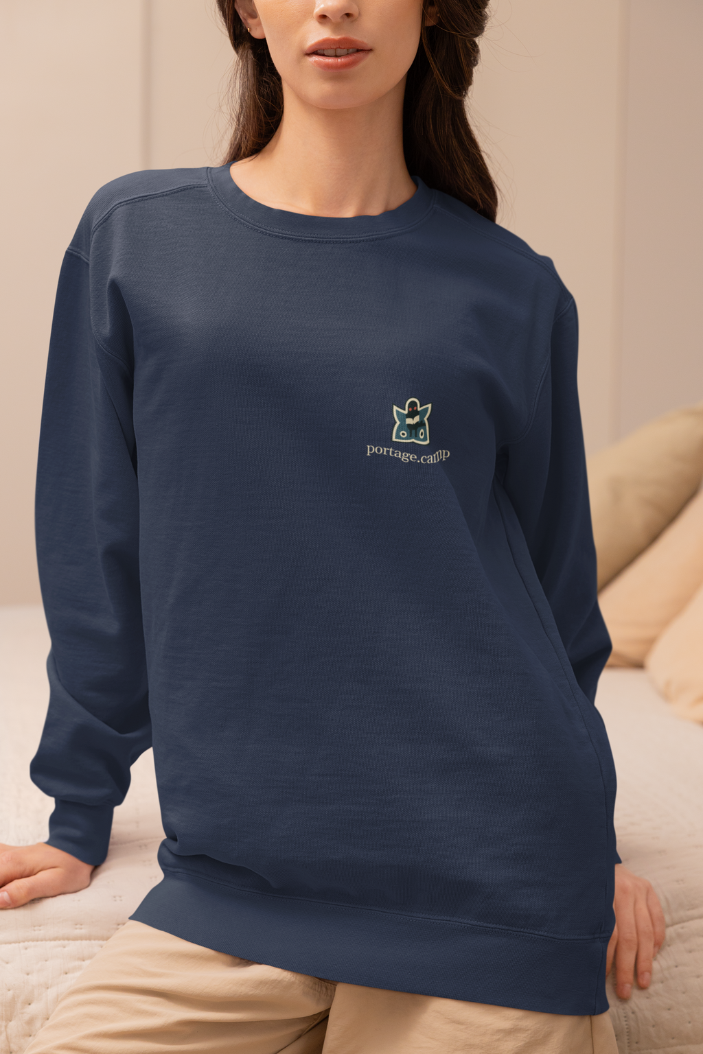 Night Library Garment-Dyed Crewneck in True Navy, worn by a woman seated on a bed at home, with the Night Library logo and portage.camp on the chest. Digital product mockup.