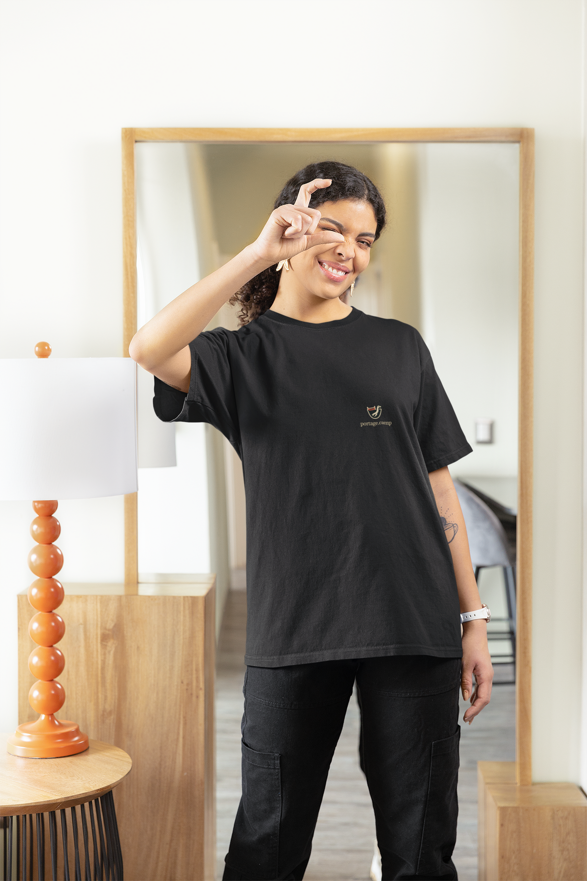Seen Briefly Garment-Dyed Tee in Black, worn by a woman at home, pretending to take a photo, with the Seen Briefly logo and portage.camp on the chest. Digital product mockup.