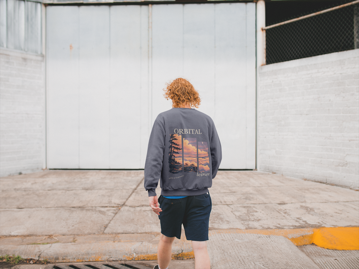 Edge of Evening Garment-Dyed Crewneck in Denim, worn by a man seen from behind walking away across an empty yard, showing the large print on the back. Digital product mockup.
