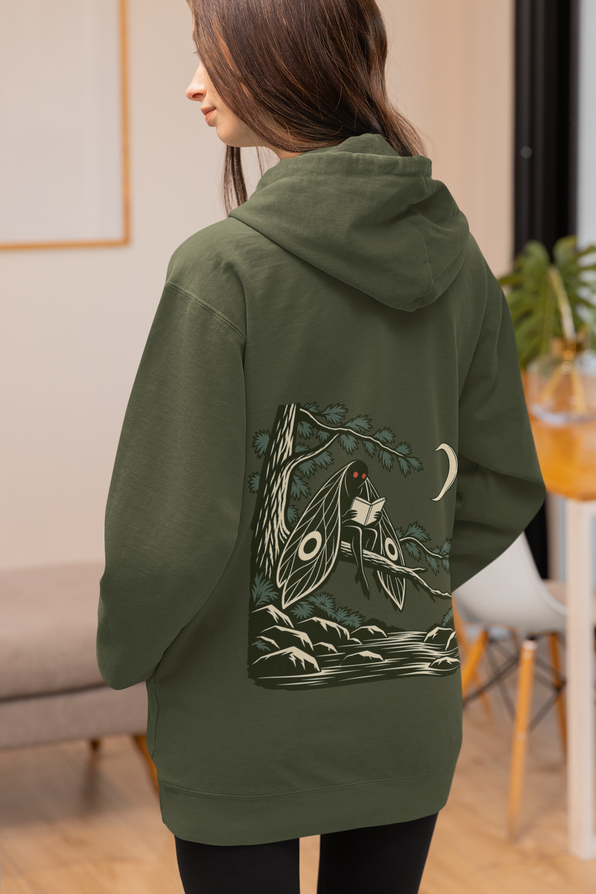 Night Library Midweight Hoodie in Army, worn by a woman seen from behind at home, showing the large print on the back. Digital product mockup.