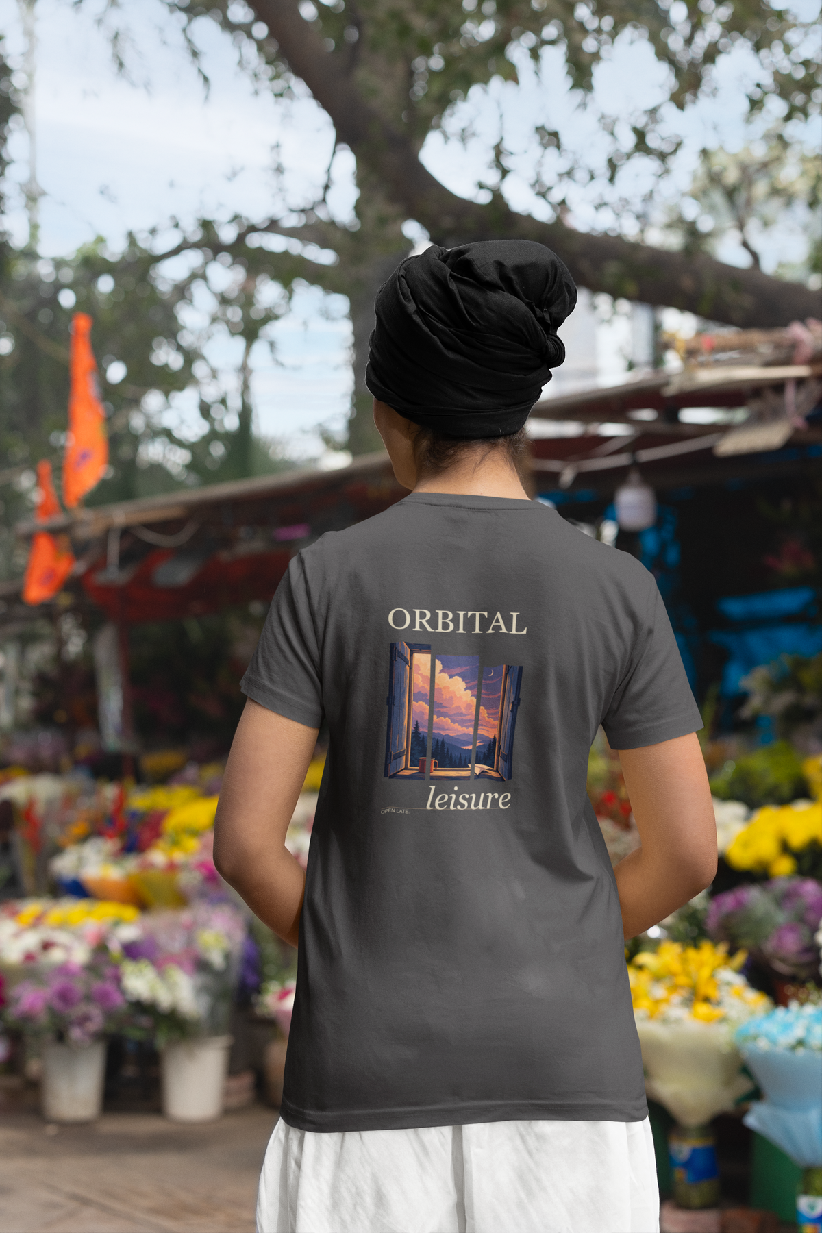 Open Late Garment-Dyed Tee in Graphite, worn by a woman seen from behind in a turban by a flower market, showing the large print on the back. Digital product mockup.