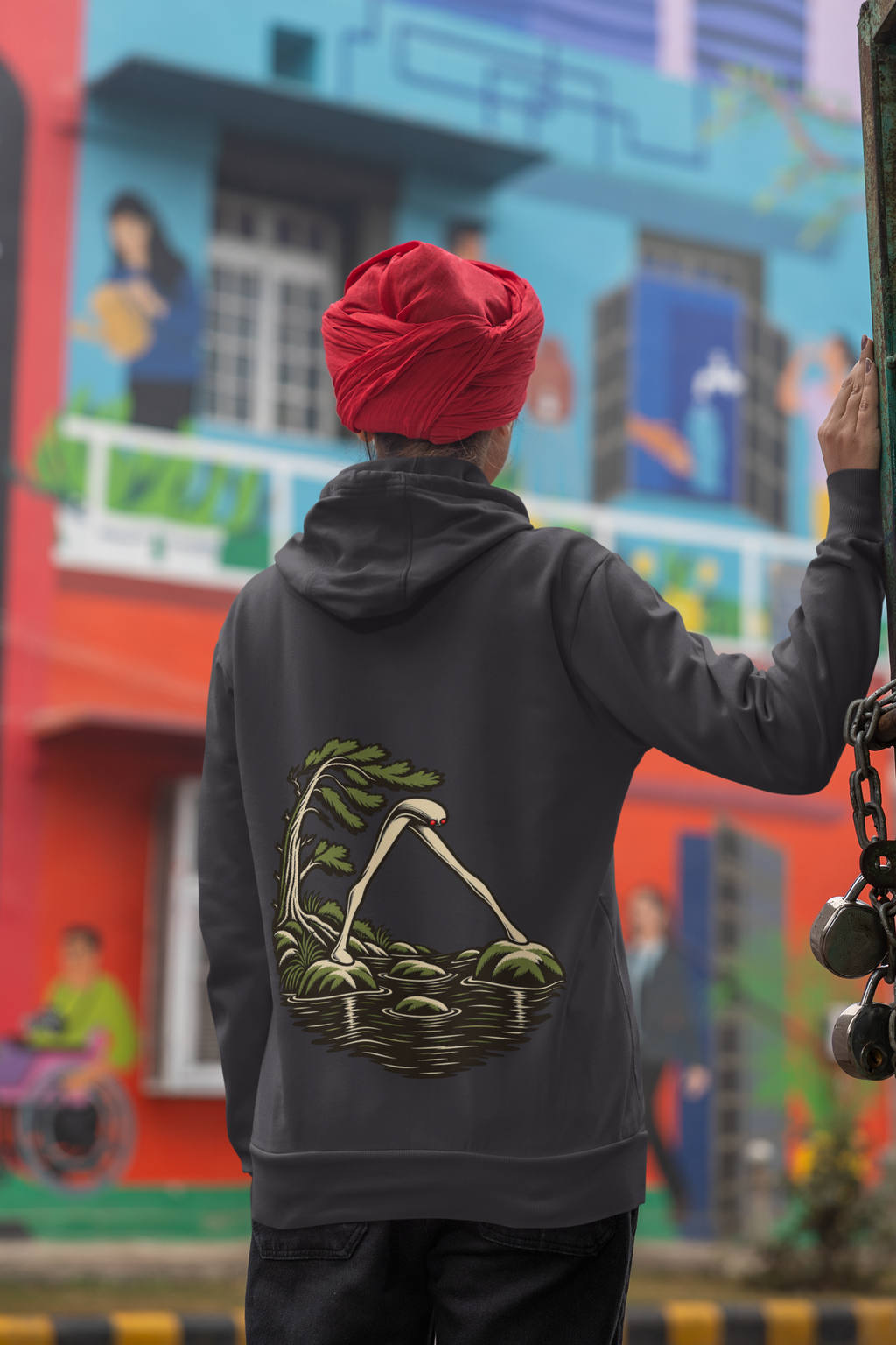 Open Late Heavyweight Hoodie in Black, worn by a woman seen from behind in a turban by a painted mural, showing the large print on the back. Digital product mockup.