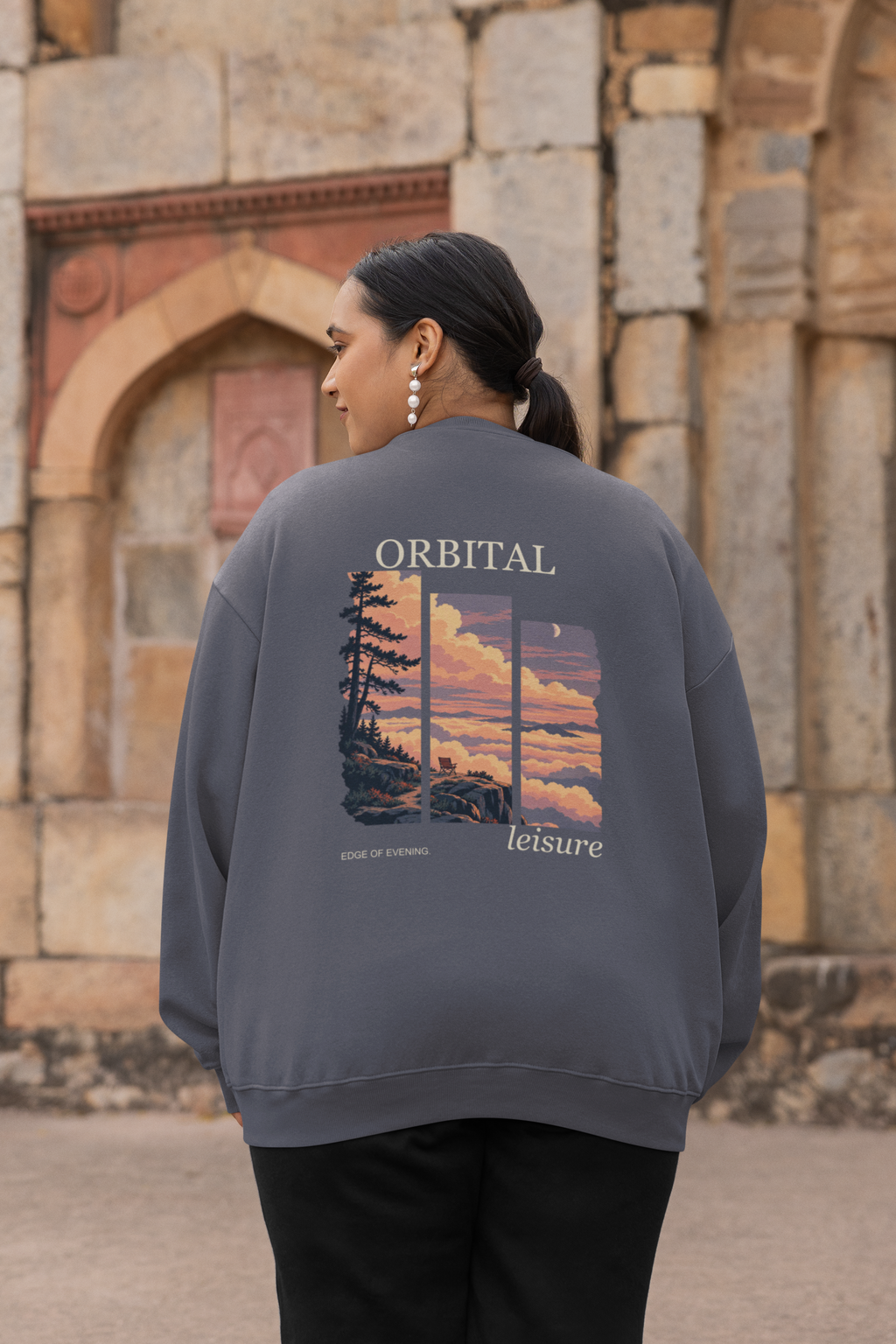 Edge of Evening Garment-Dyed Crewneck in Denim, worn by a woman seen from behind by an old stone building, showing the large print on the back. Digital product mockup.