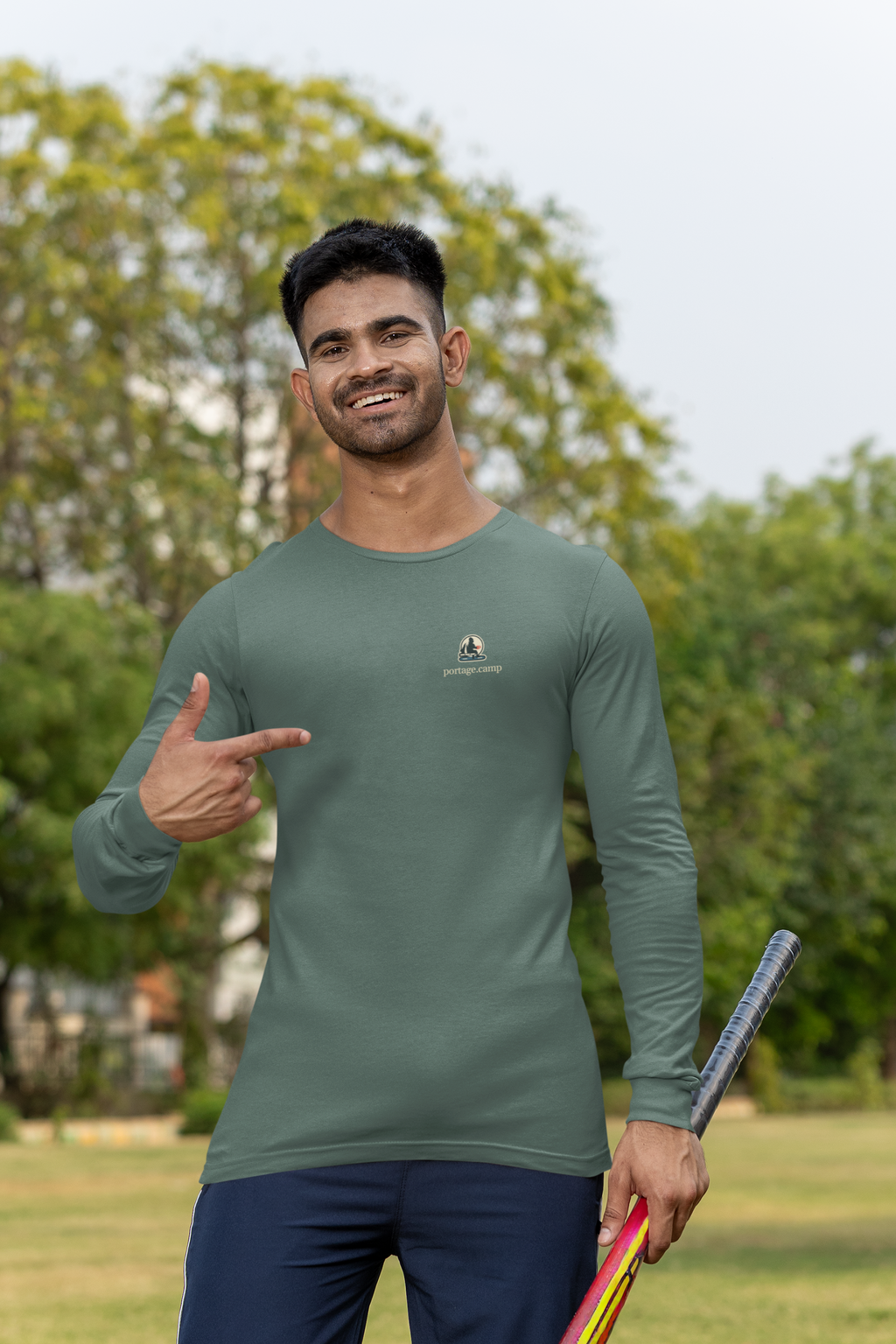 Quiet Hours Garment-Dyed Long-Sleeve Tee in Blue Spruce, worn by a man at a cricket field, with the Quiet Hours logo and portage.camp on the chest. Digital product mockup.
