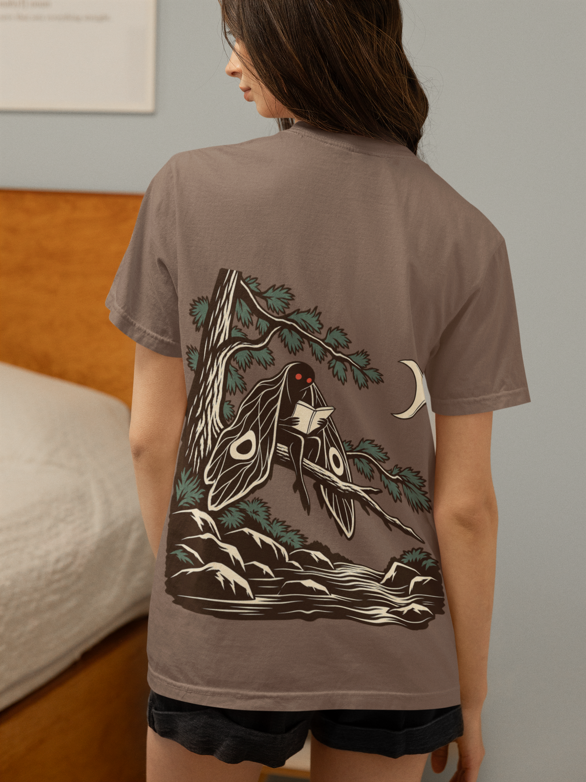 Night Library Garment-Dyed Tee in Espresso, worn by a woman seen from behind in a bedroom at home, showing the large print on the back. Digital product mockup.