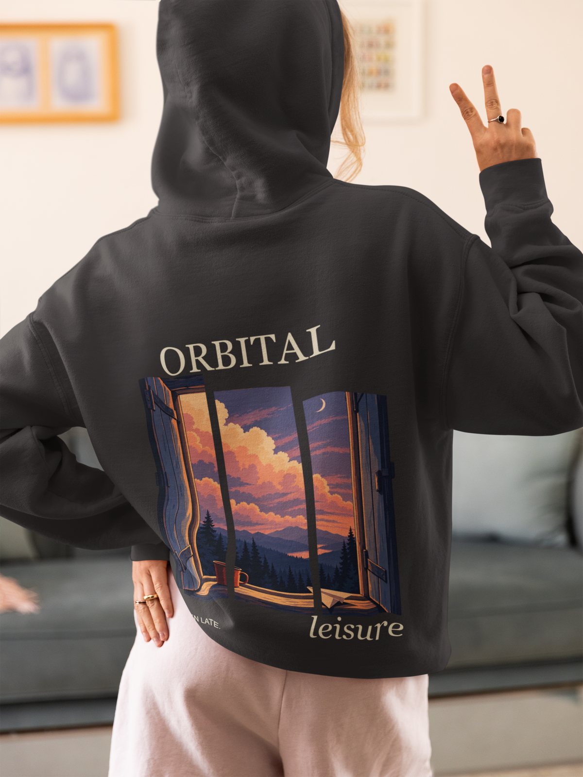 Cloud Ferry Heavyweight Hoodie in Black, worn by a woman seen from behind at home, making a peace sign, showing the large print on the back. Digital product mockup.