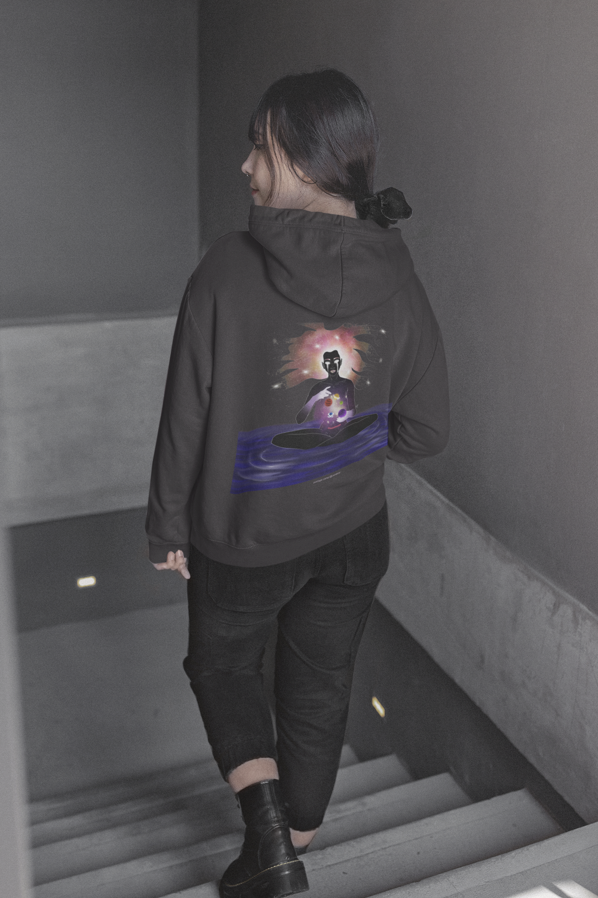 Orbit Keeper Midweight Hoodie in Black, worn by a woman seen from behind walking down a staircase, showing the large print on the back. Digital product mockup.