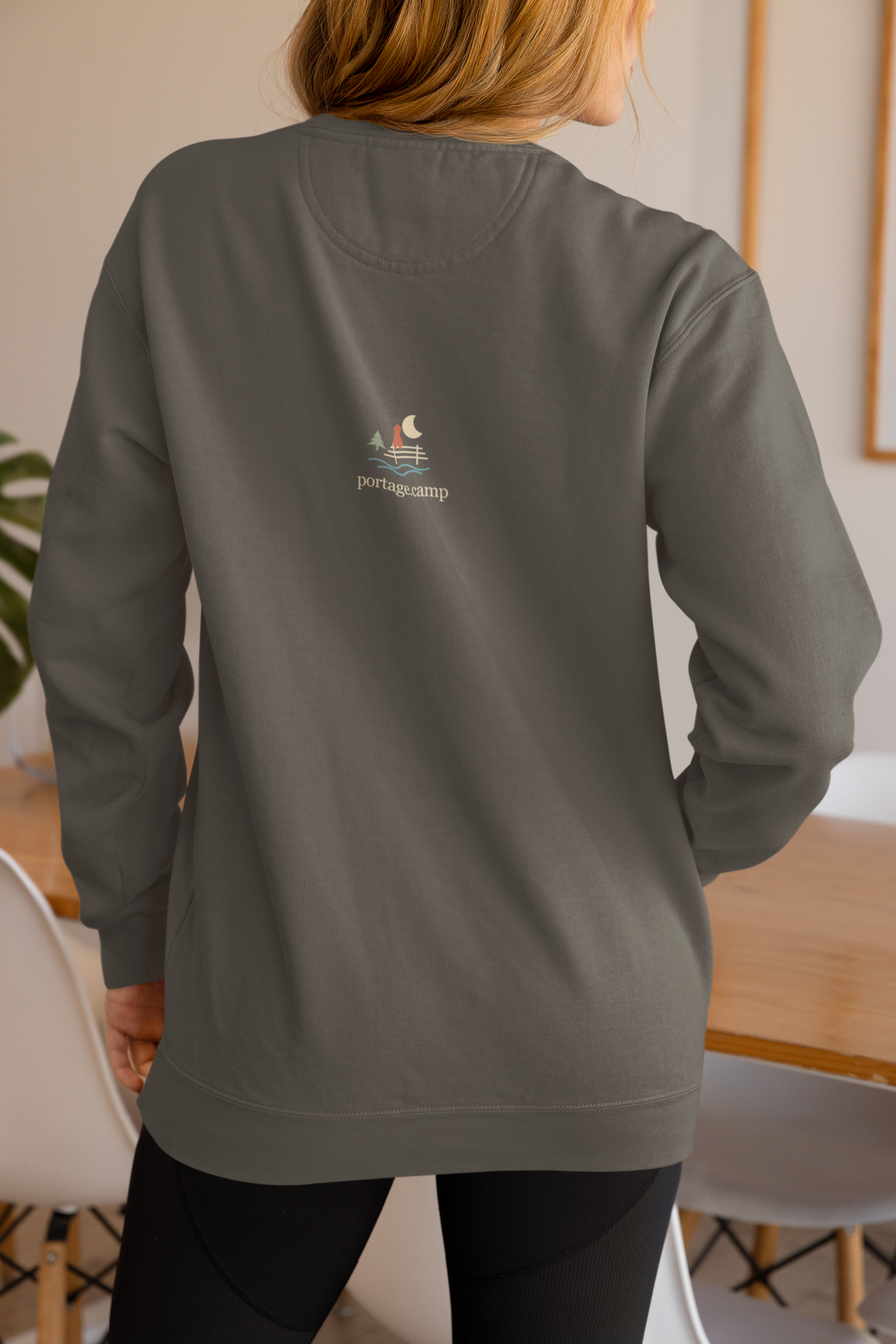Quiet Hours Garment-Dyed Crewneck (Front Print) in Pepper, worn by a woman seen from behind at home, showing the portage.camp emblem at the upper back. Digital product mockup.