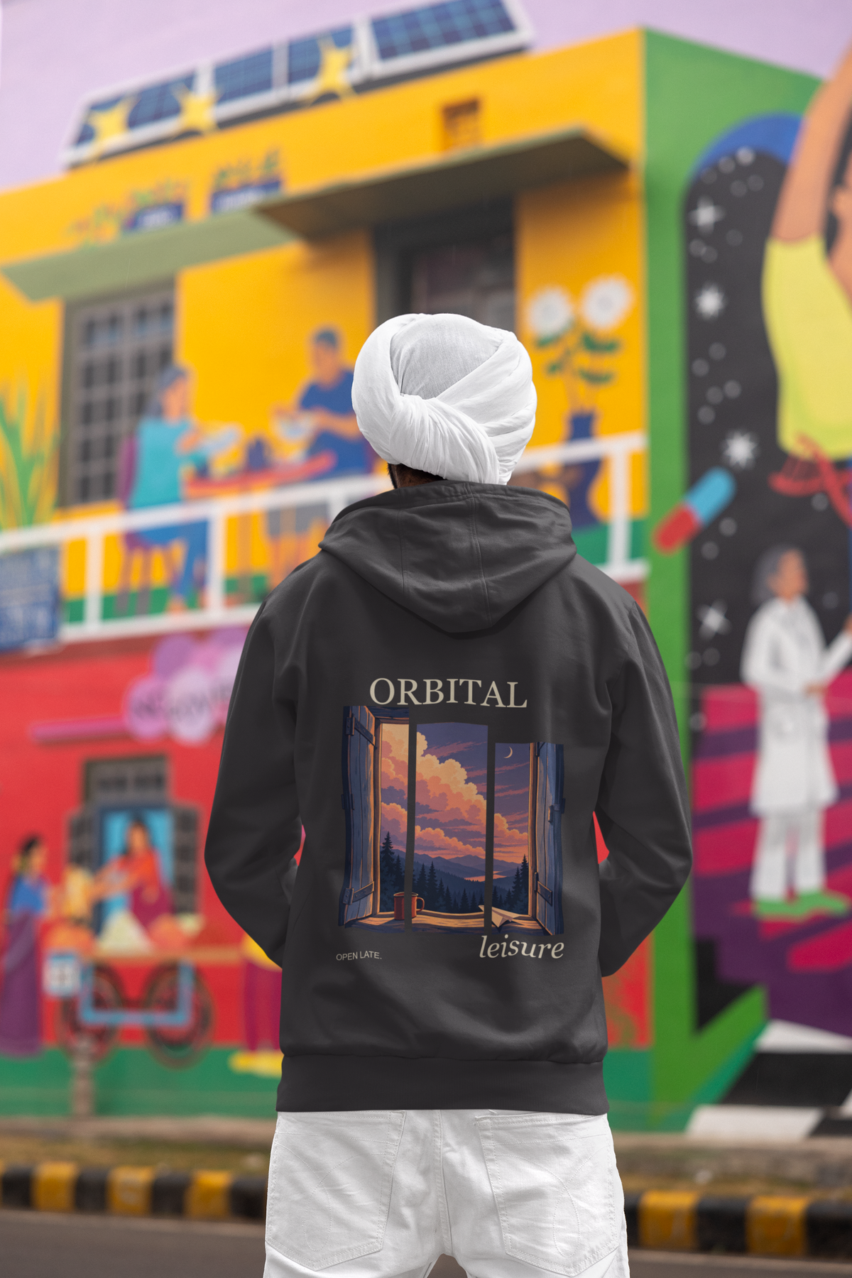 Open Late Heavyweight Hoodie in Black, worn by a man seen from behind in a turban by a painted mural, showing the large print on the back. Digital product mockup.